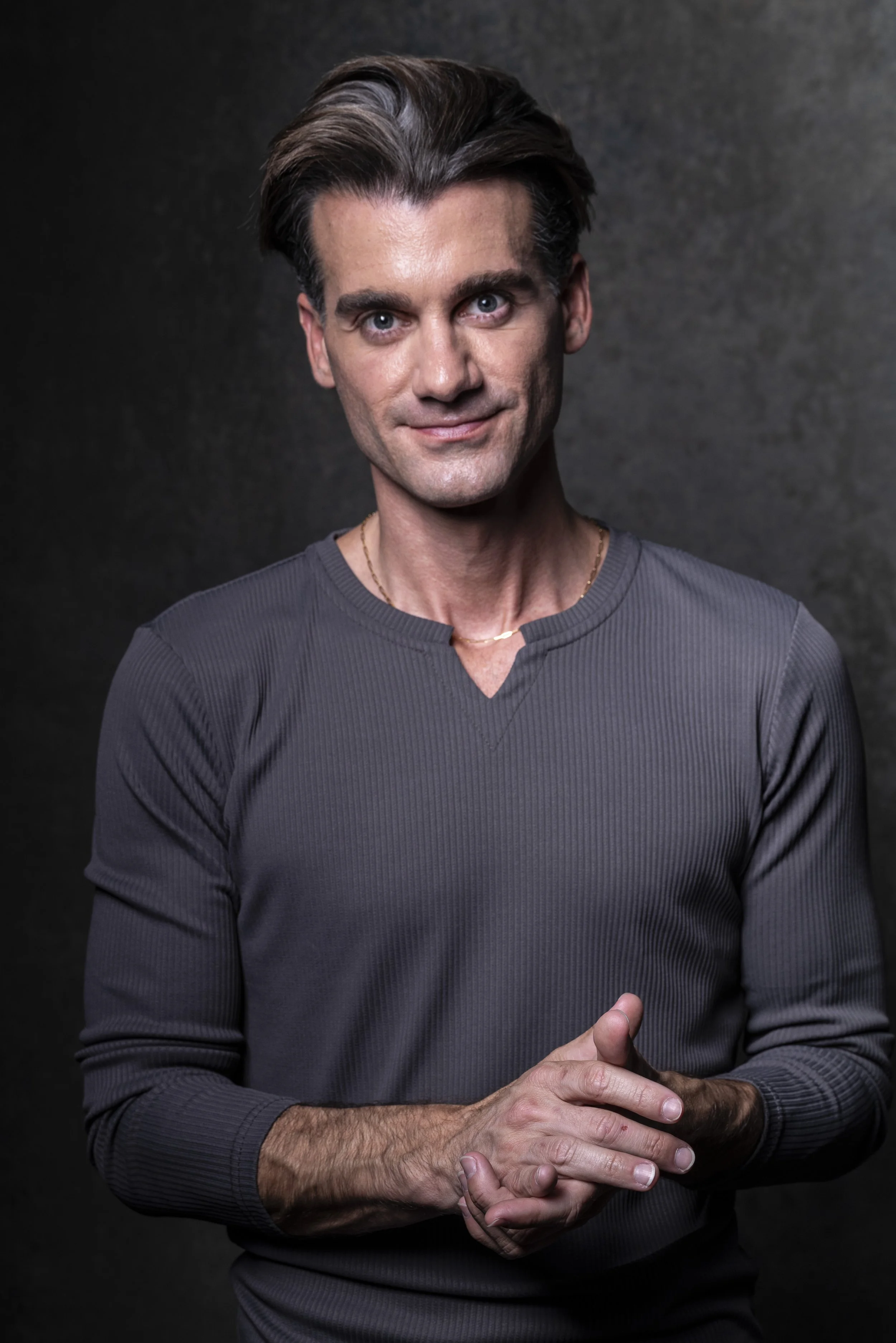 A man with dark hair and blue eyes wearing a gray long-sleeve shirt and gold necklace, standing against a dark background.