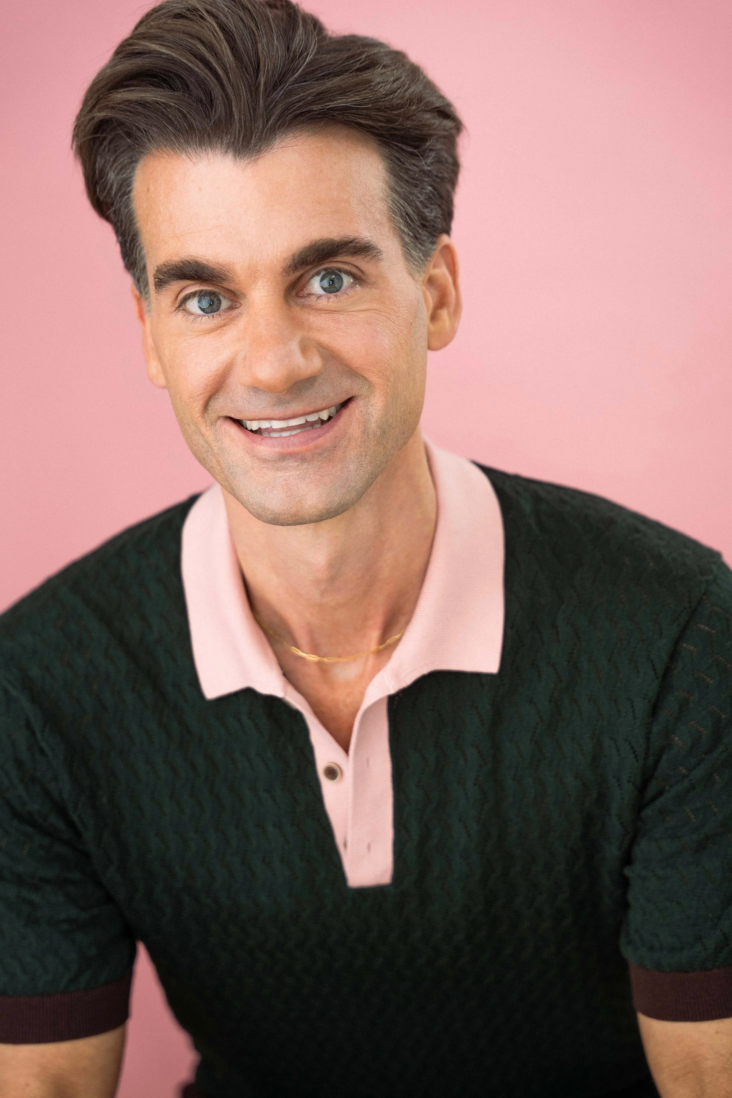 A smiling man with brown hair and blue eyes, wearing a pink collared shirt and a dark green sweater with maroon cuffs, against a pink background.