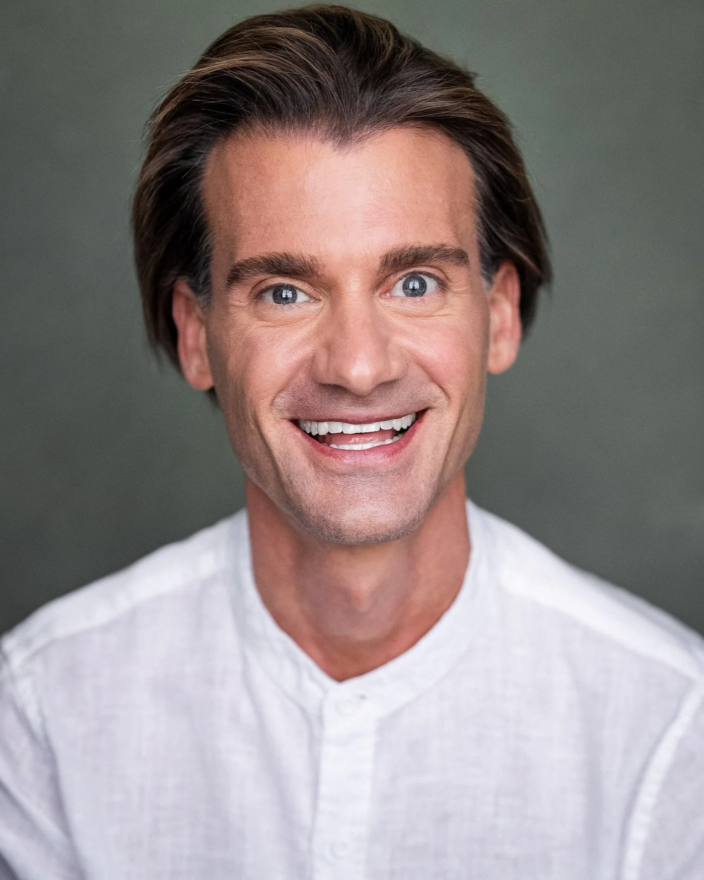 A man with short brown hair and blue eyes smiling, wearing a light-colored shirt against a plain background.