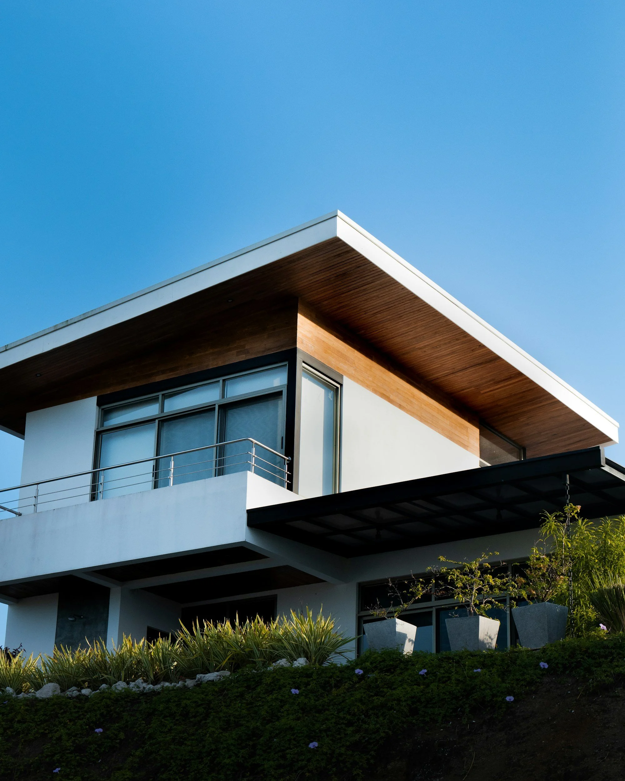 Modern multi-story house with large windows, wood accents, and a balcony, surrounded by landscaped garden with plants and flowers, under a clear blue sky.