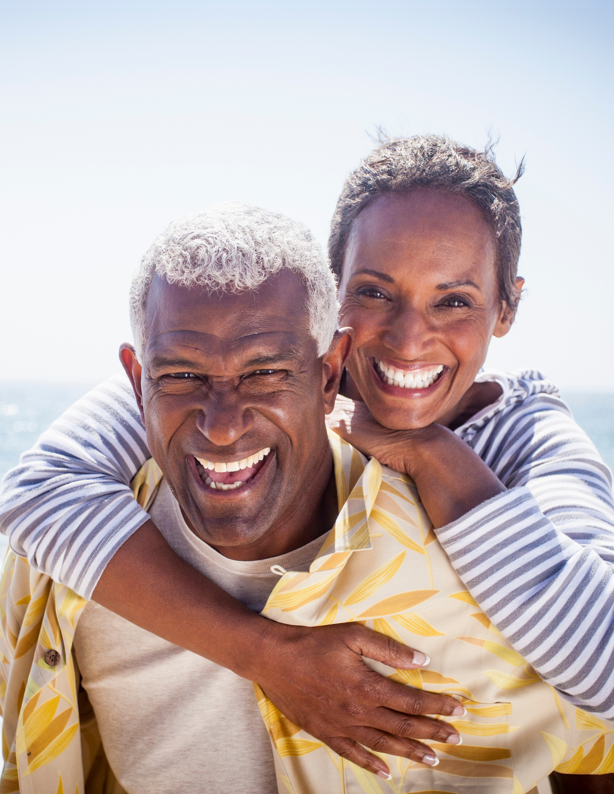 Close-up of an elderly couple smiling and embracing near the beach on a sunny day, woman with short gray hair, man with curly gray hair, wearing striped clothing.