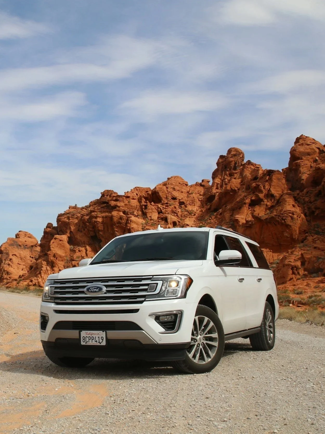 A white Ford SUV parked on a dirt road in a desert landscape with red rock formations and a blue sky with scattered clouds.