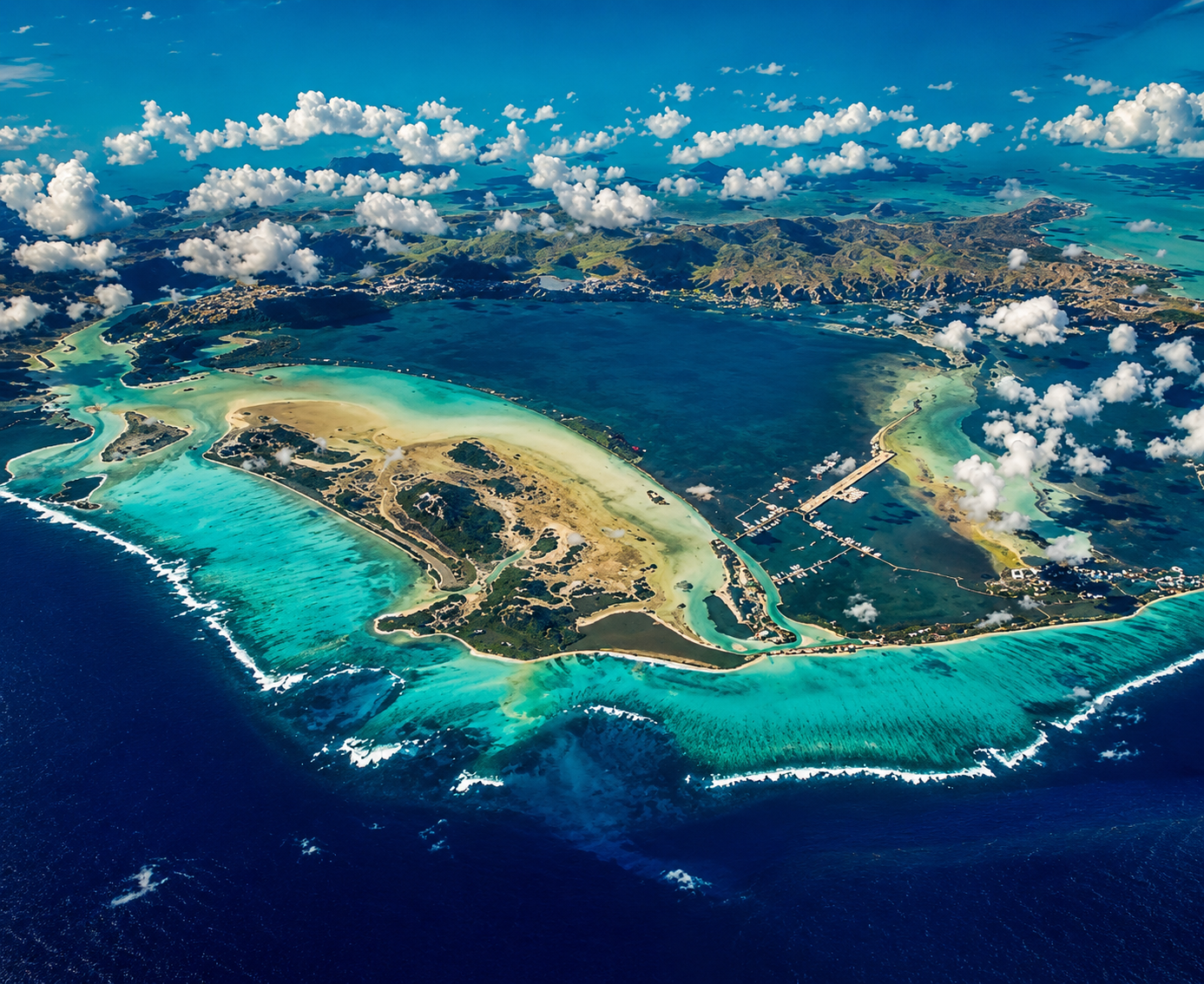 Vue aérienne d'une île entourée d'eau turquoise, avec des plages, des routes et quelques bâtiments, sous un ciel partiellement nuageux.