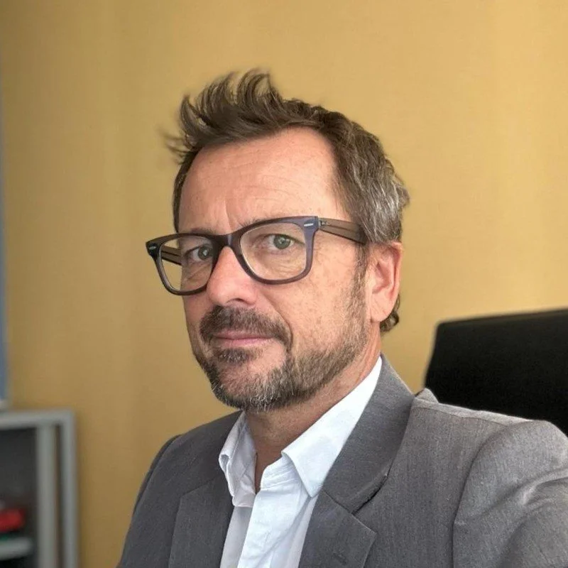 Un homme avec des lunettes, une veste grise et une chemise blanche pose pour une photo dans un bureau, avec un mur jaune en arrière-plan.