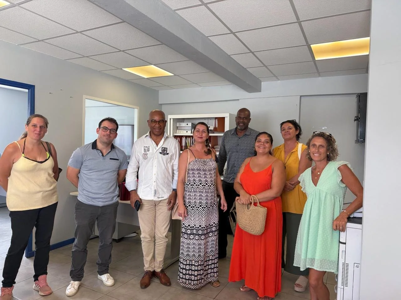 Groupe de neuf personnes posant en intérieur, dans une pièce avec plafond en dalles suspendues et éclairage encastré, certains tenant des sacs ou appareils électroniques.