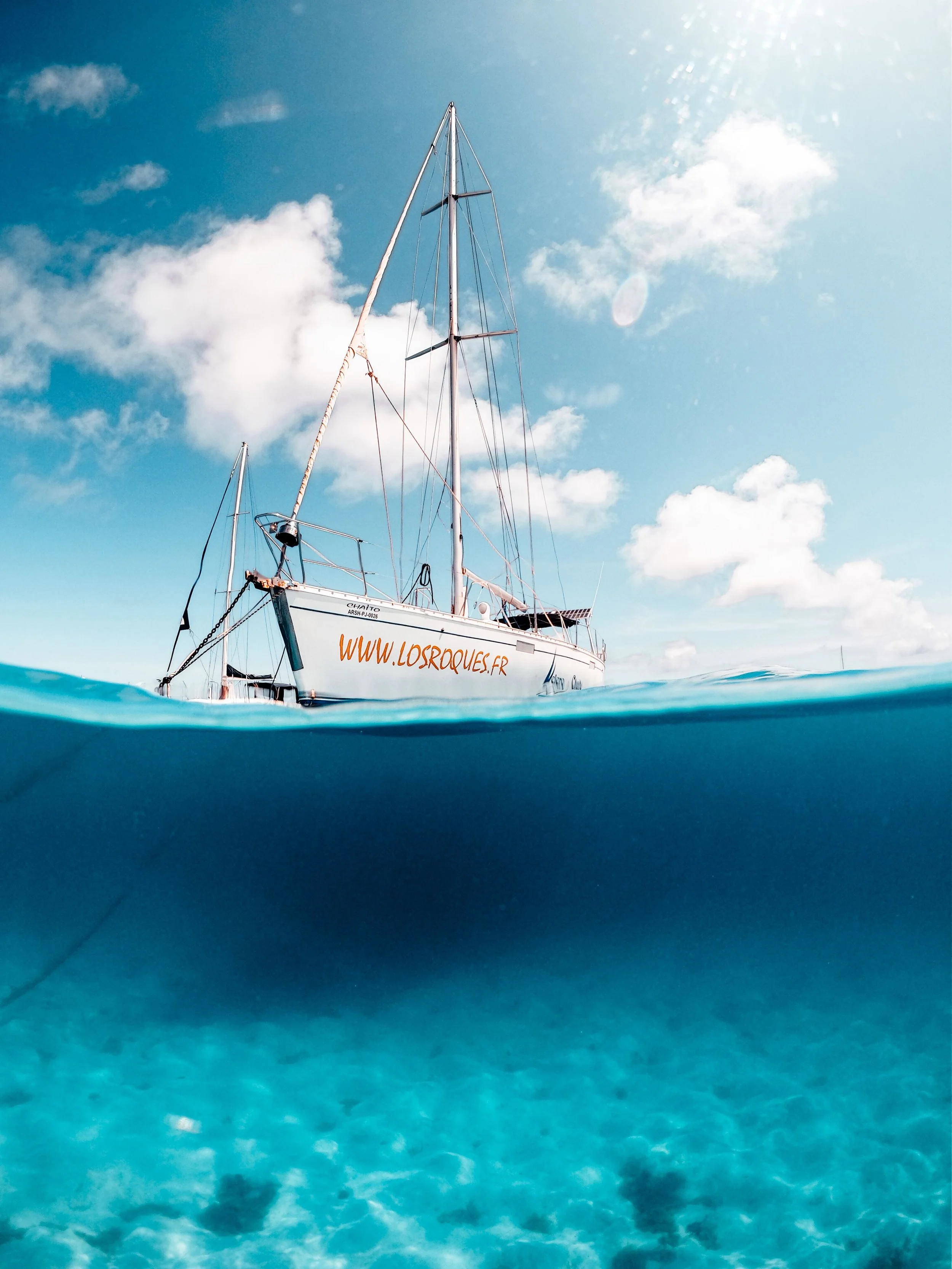 Voile de bateau vue partiellement sous l'eau avec un ciel bleu, quelques nuages et soleil éclatant.