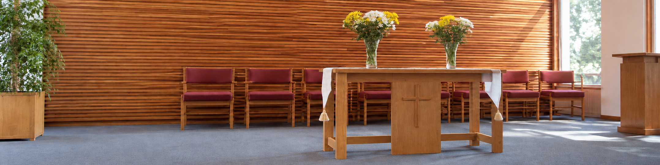 An altar in a church with a wooden background, decorated with two vases of yellow and white flowers, and surrounded by several red-cushioned chairs.