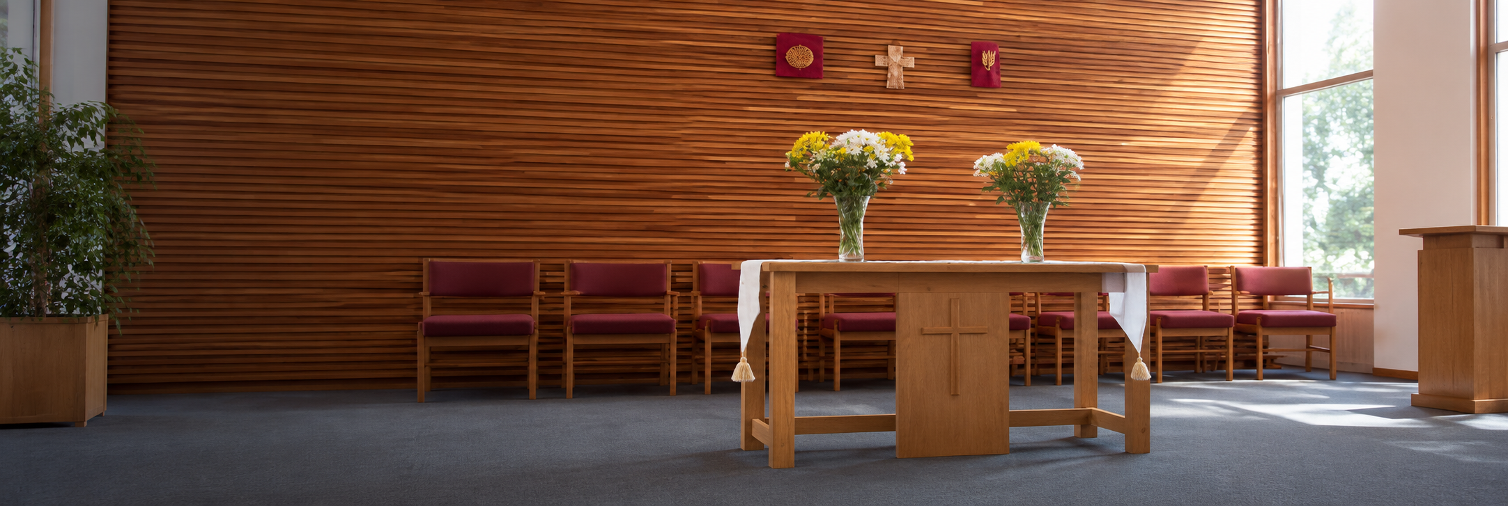Inside a church, a wooden altar with two large flower vases filled with yellow and white flowers, a wooden cross on the front, burgundy chairs against a wooden wall, and three Christian symbols hanging on the wall above the altar. Sunlight streams in through large windows.