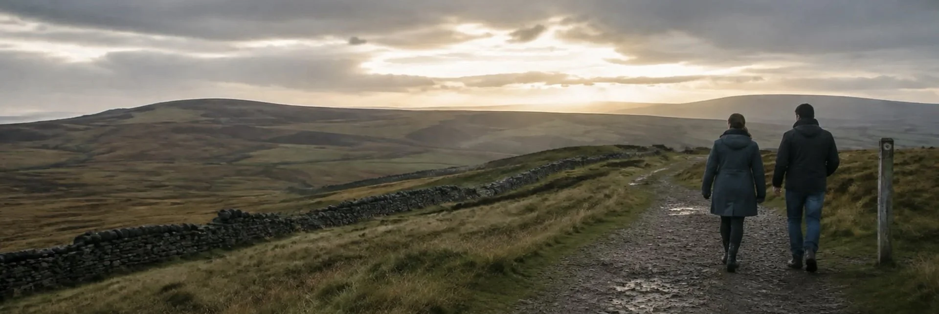A couple walking along a gravel path in a hilly, rural landscape during sunset, with cloudy skies and a low stone wall on the left.