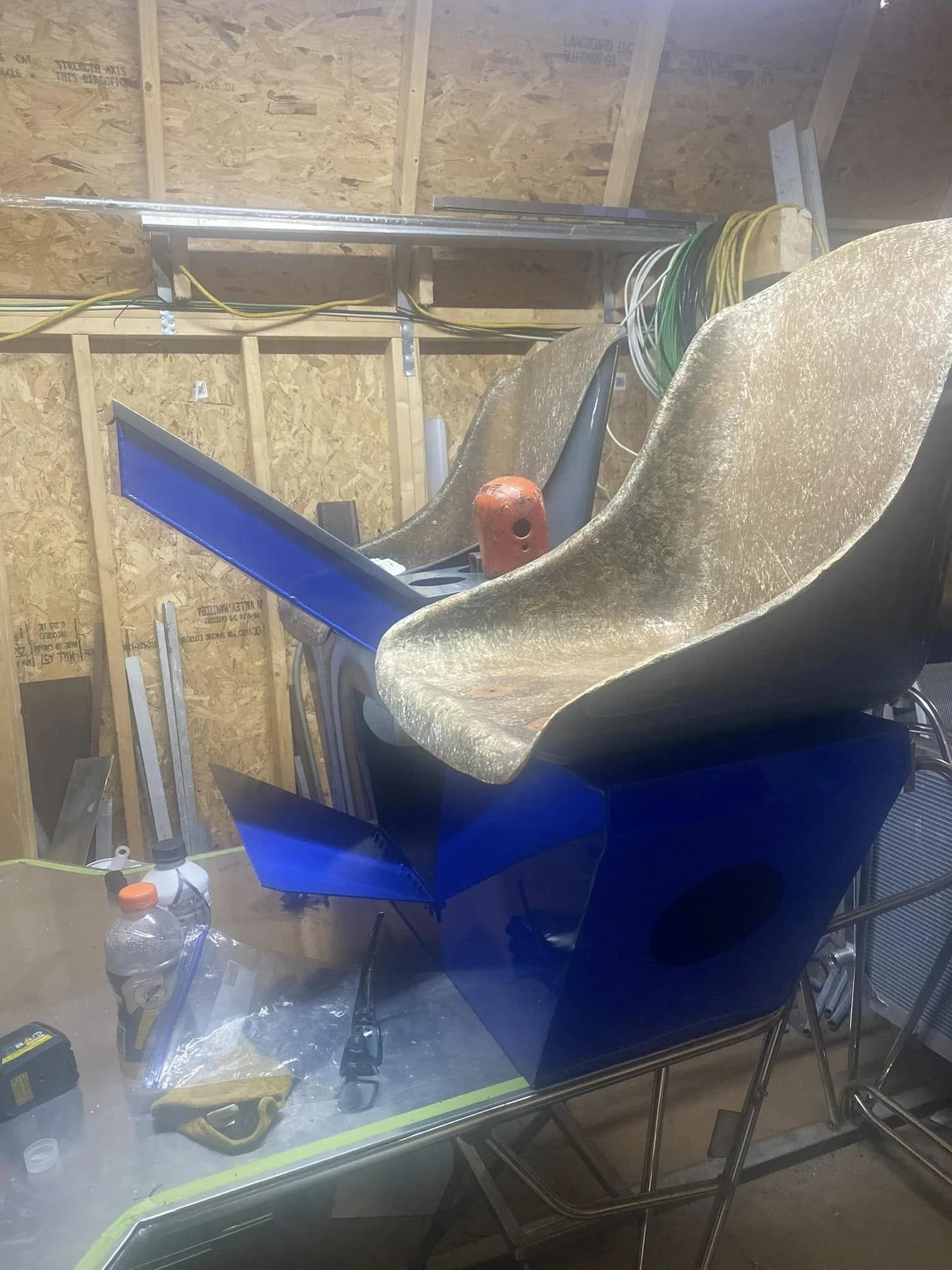 Photo of a blue motorcycle or scooter inside a workshop or garage, with a gold-colored fairing, partially visible. The background features wooden wall framing and electrical wires.