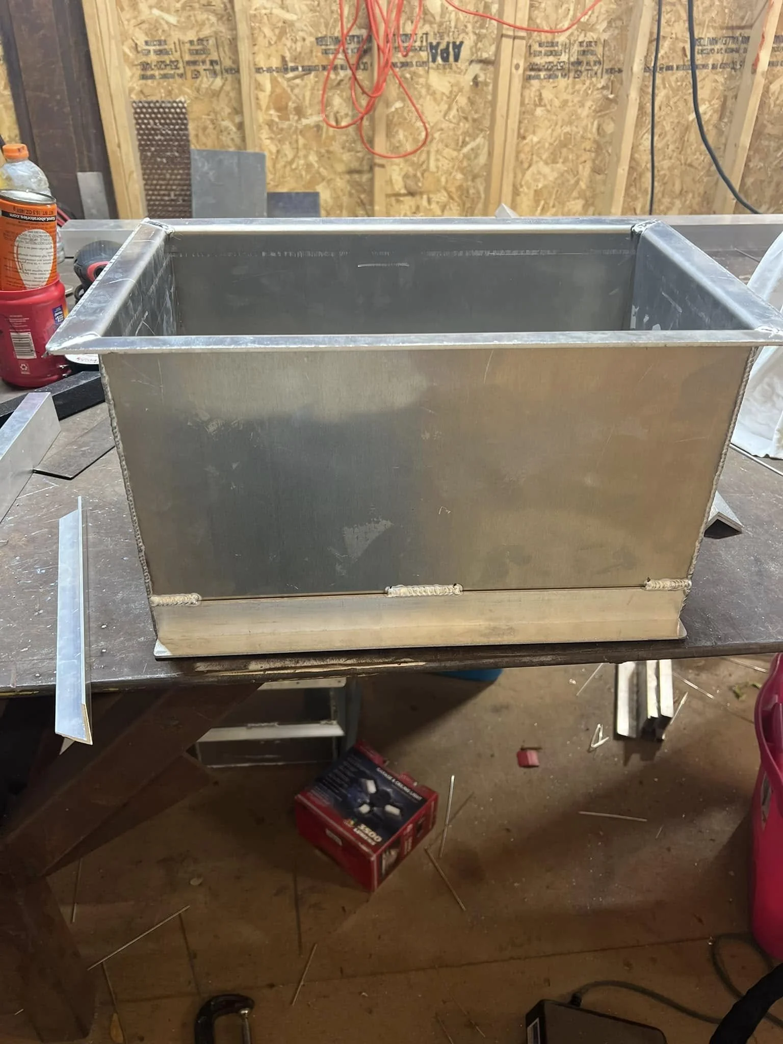 A metal rectangular container on a workbench in a workshop, surrounded by tools and supplies, with a wooden wall and construction materials in the background.