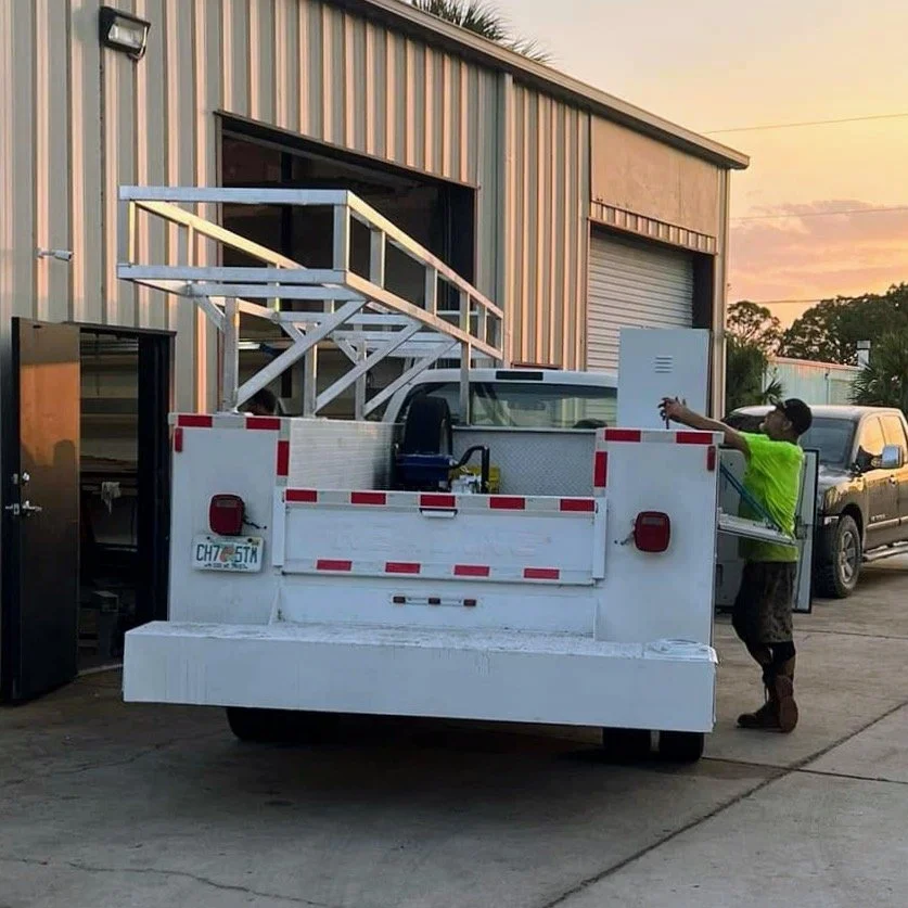 A worker in a neon green shirt loading tools into a white utility truck parked outside a beige industrial building during sunset.