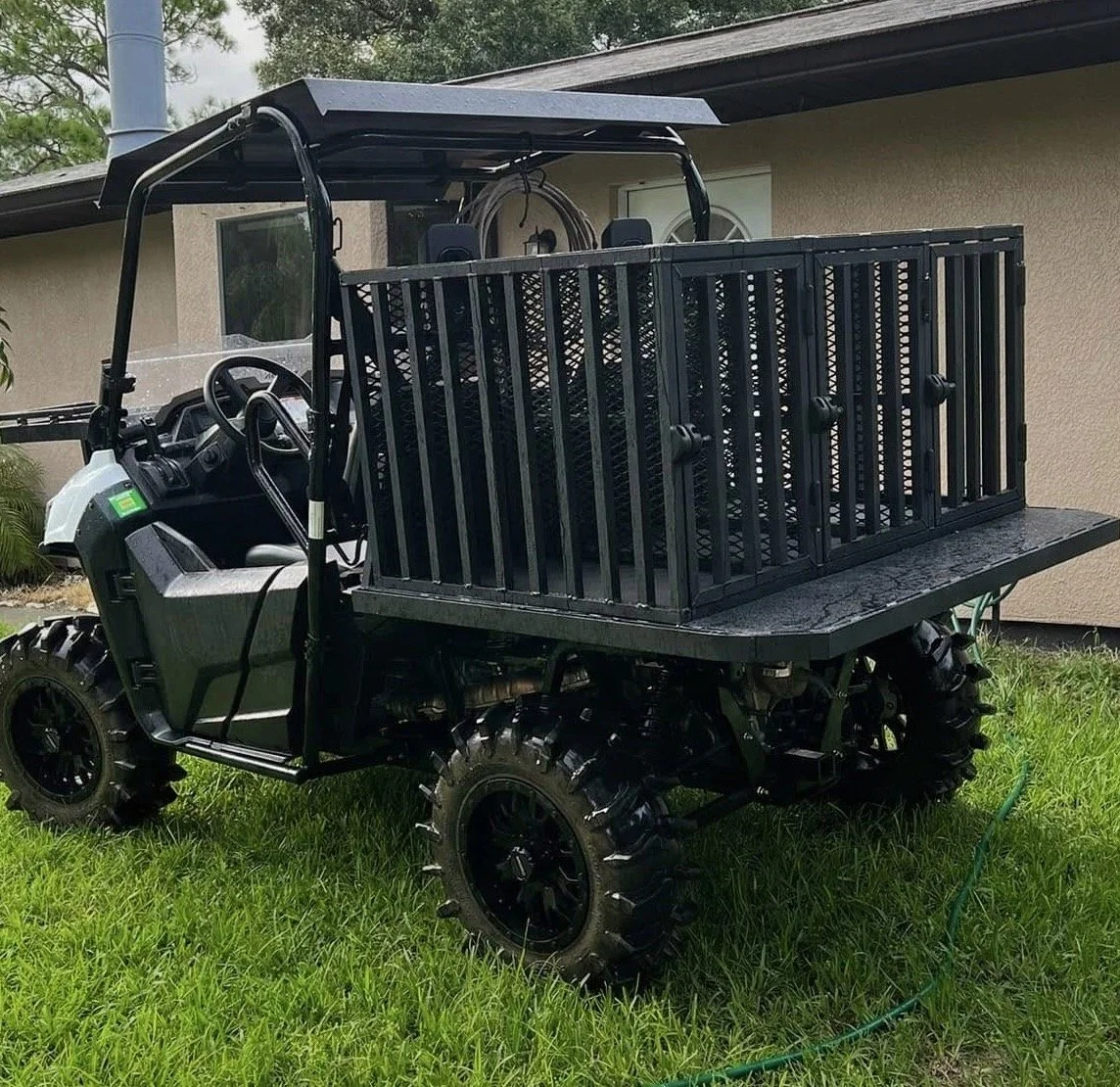 Black utility vehicle with large off-road tires and a cargo bed containing black plastic crates, parked on a grassy area near a house.
