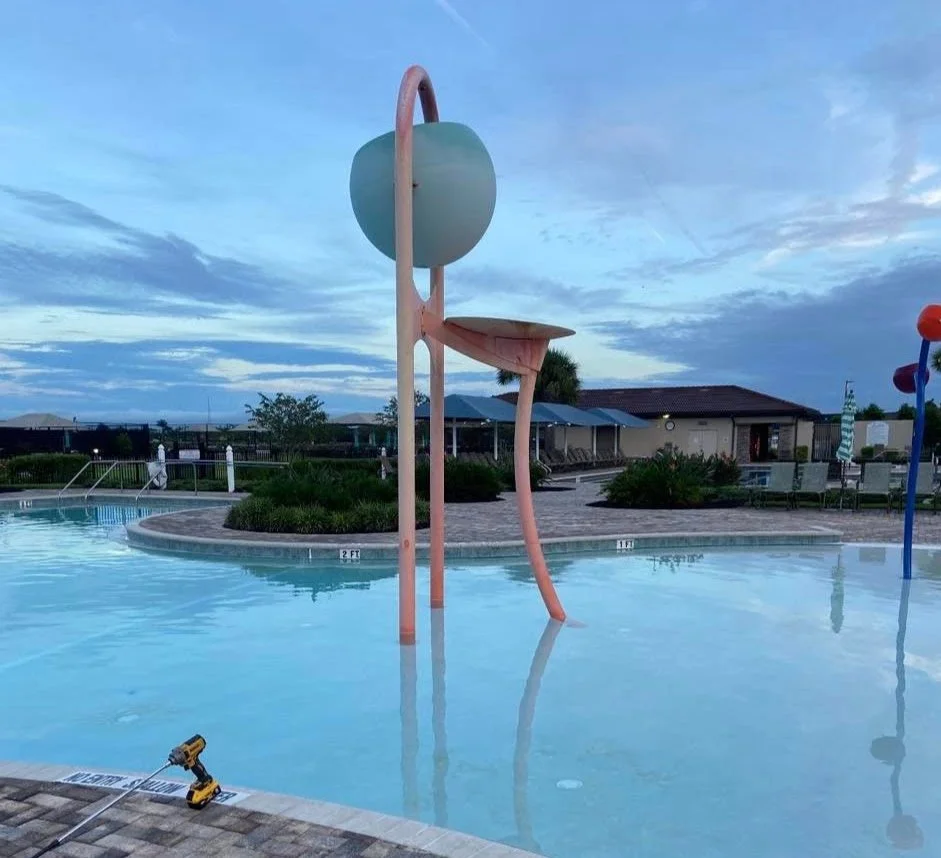 Empty swimming pool with colorful water spray structures and a cloudy sky.