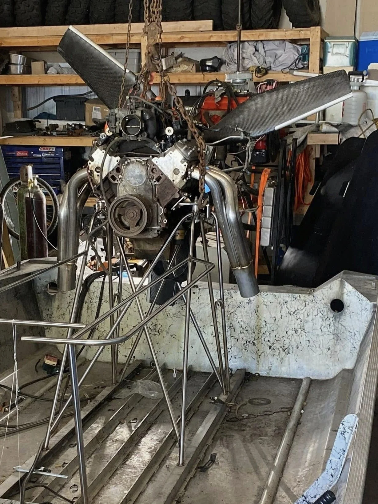 A car engine is suspended above a bathtub, likely in a garage or workshop, with metal rods and chains holding it in place, surrounded by tools and workshop supplies.