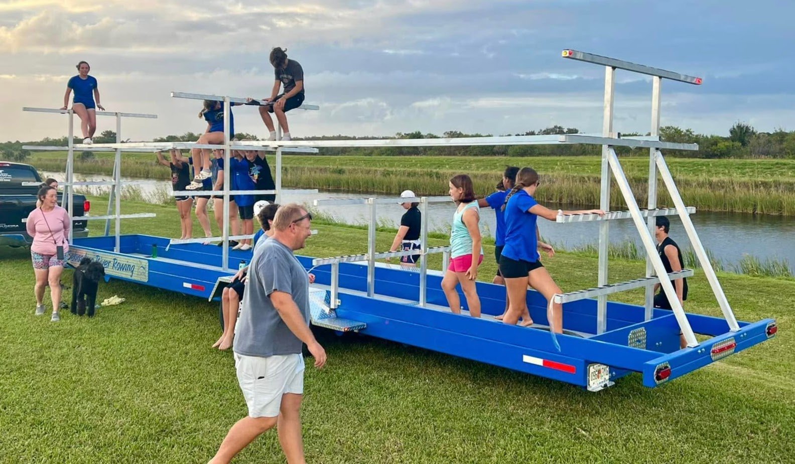 People preparing for a rowing practice on a specialized floating rig outdoors near a water body, with grassy fields and cloudy sky in the background.