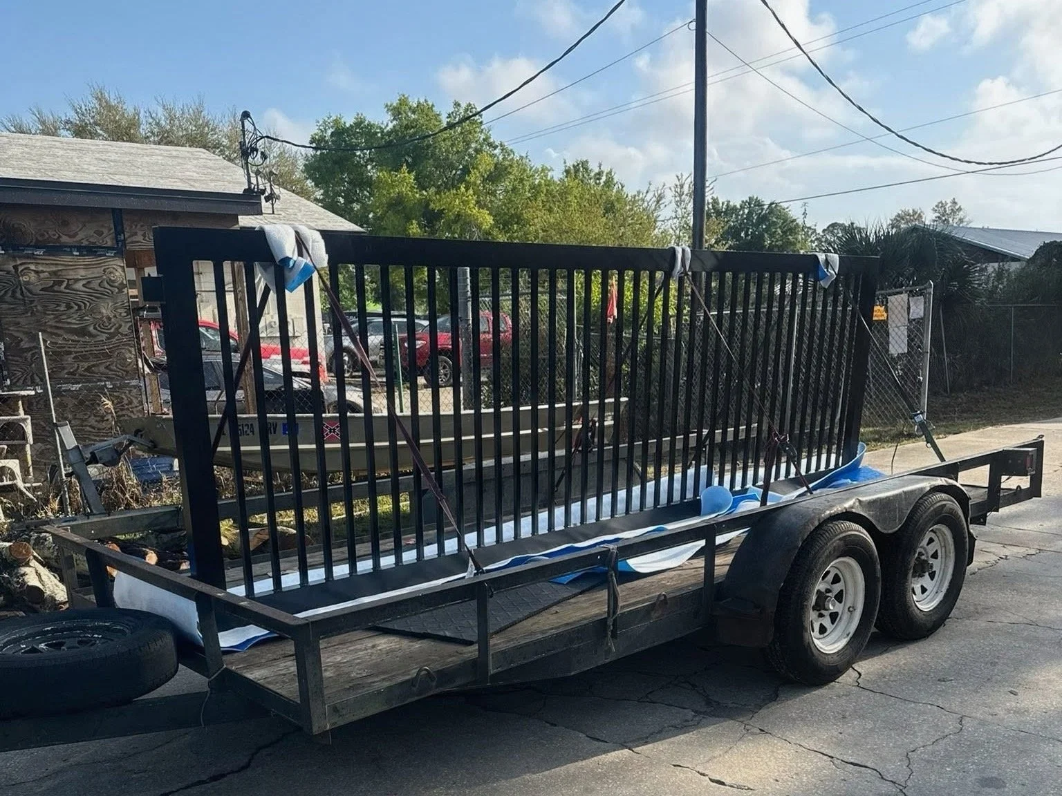 A trailer with blue and white balloons tied to the metal bars, parked on a cracked asphalt surface outdoors near a small shed and trees.