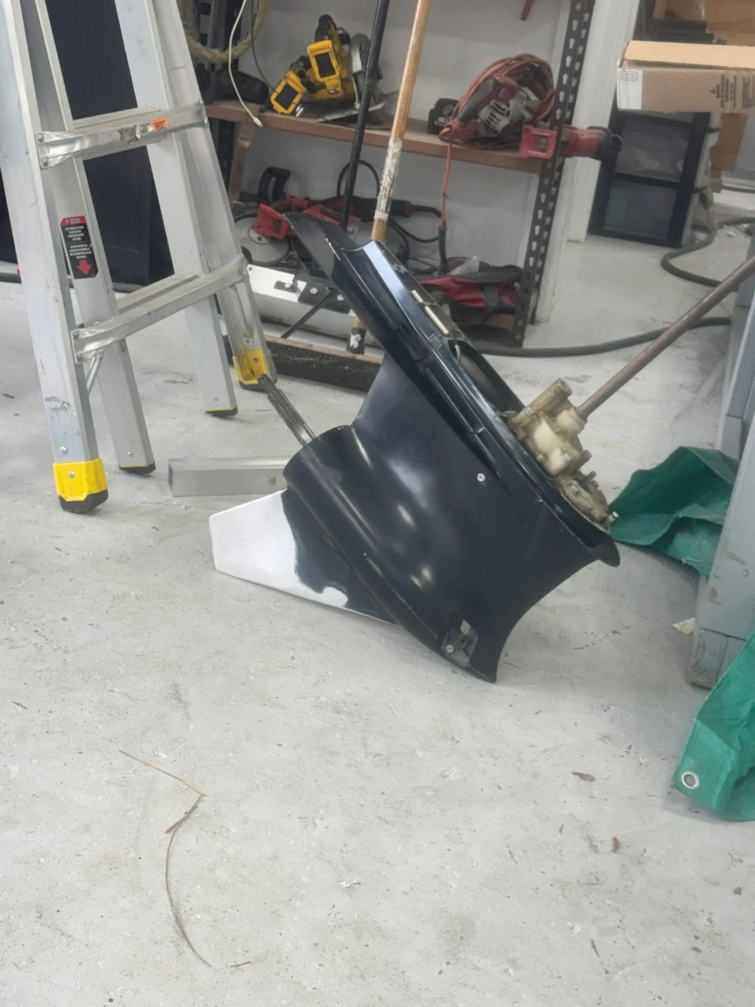 A cluttered workshop with a metal ladder, power tools, and construction materials scattered on a concrete floor.
