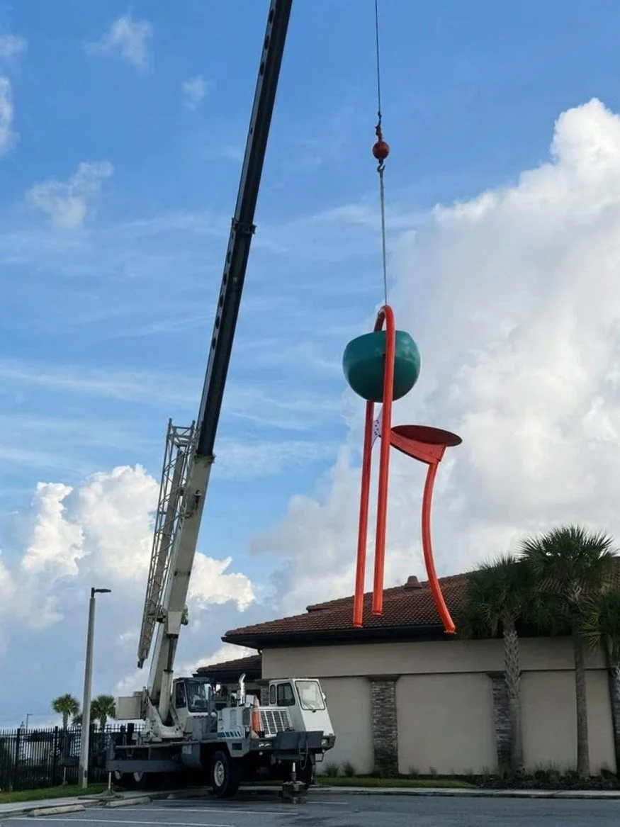 A crane lifting a large, modern sculpture of a chair with a ball on it, placed on top of a building with a tiled roof. There are palm trees and a partly cloudy sky in the background.