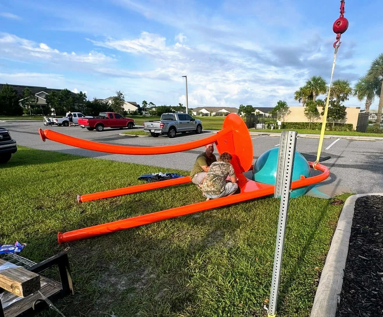 Two men working on a large disassembled playground swing set on the grass near a parking lot.