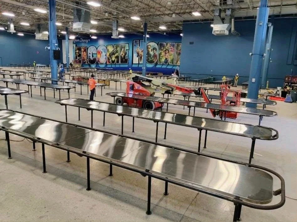 An indoor space under construction with long metallic tables, scaffolding equipment, and workers setting up. The wall features large letters spelling 'WELCOME' with Disney-themed artwork.