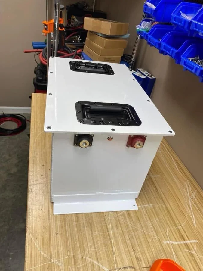 A white electrical or electronic device with black handles and gold and red connectors on a wooden workbench in a workshop or lab setting.