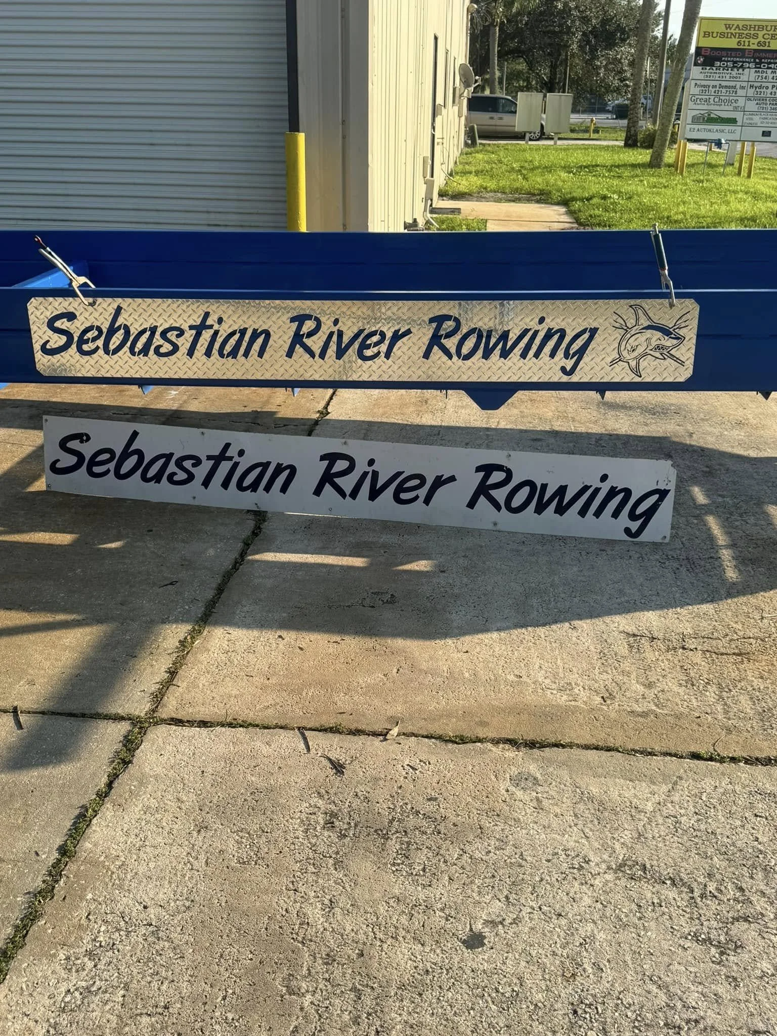 Outdoor scene with two signs for Sebastian River Rowing, one with a fish logo, placed on a sidewalk in front of a blue fence, with a building and grassy area with trees in the background.