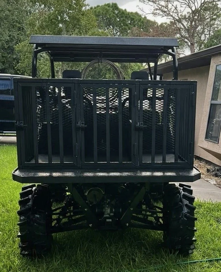 A black all-terrain vehicle with a storage cargo area at the back, parked on grass near a house. The vehicle has large, rugged tires and a canopy overhead.
