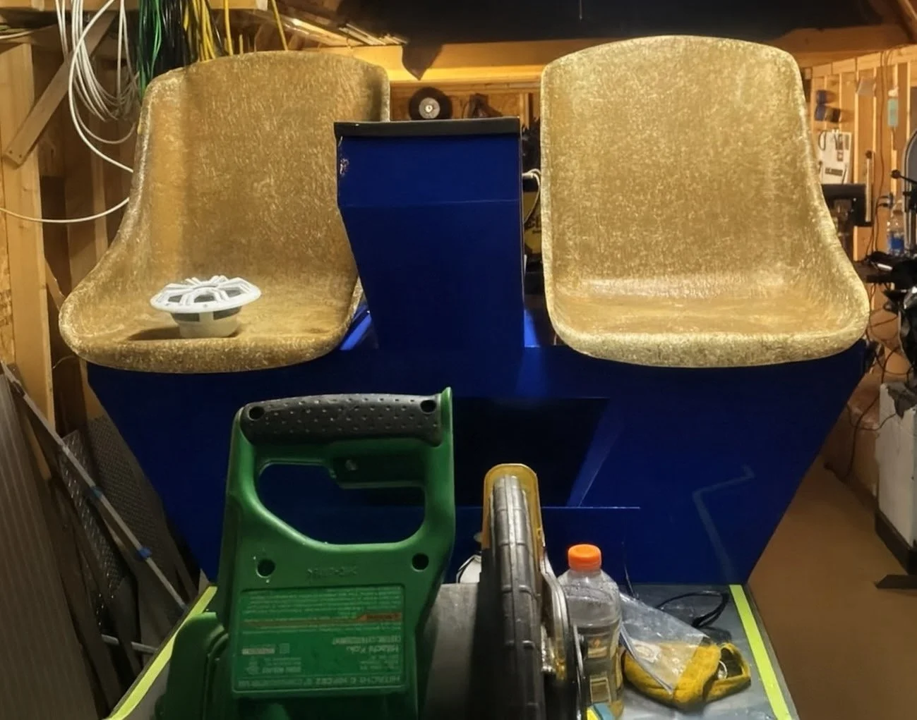 Two gold-colored velvet seats mounted on a blue structure, with a white circular component on the left seat. The setup is in a workshop with tools and equipment, including a green power saw, a water bottle, and various cords and wires.