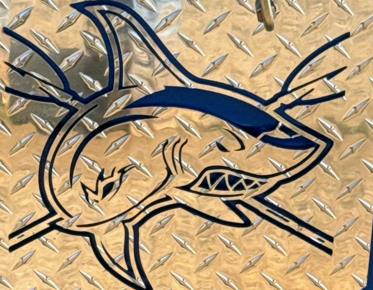 A stylized shark logo with sharp teeth and a prominent dorsal fin, painted on a metallic diamond plate surface.