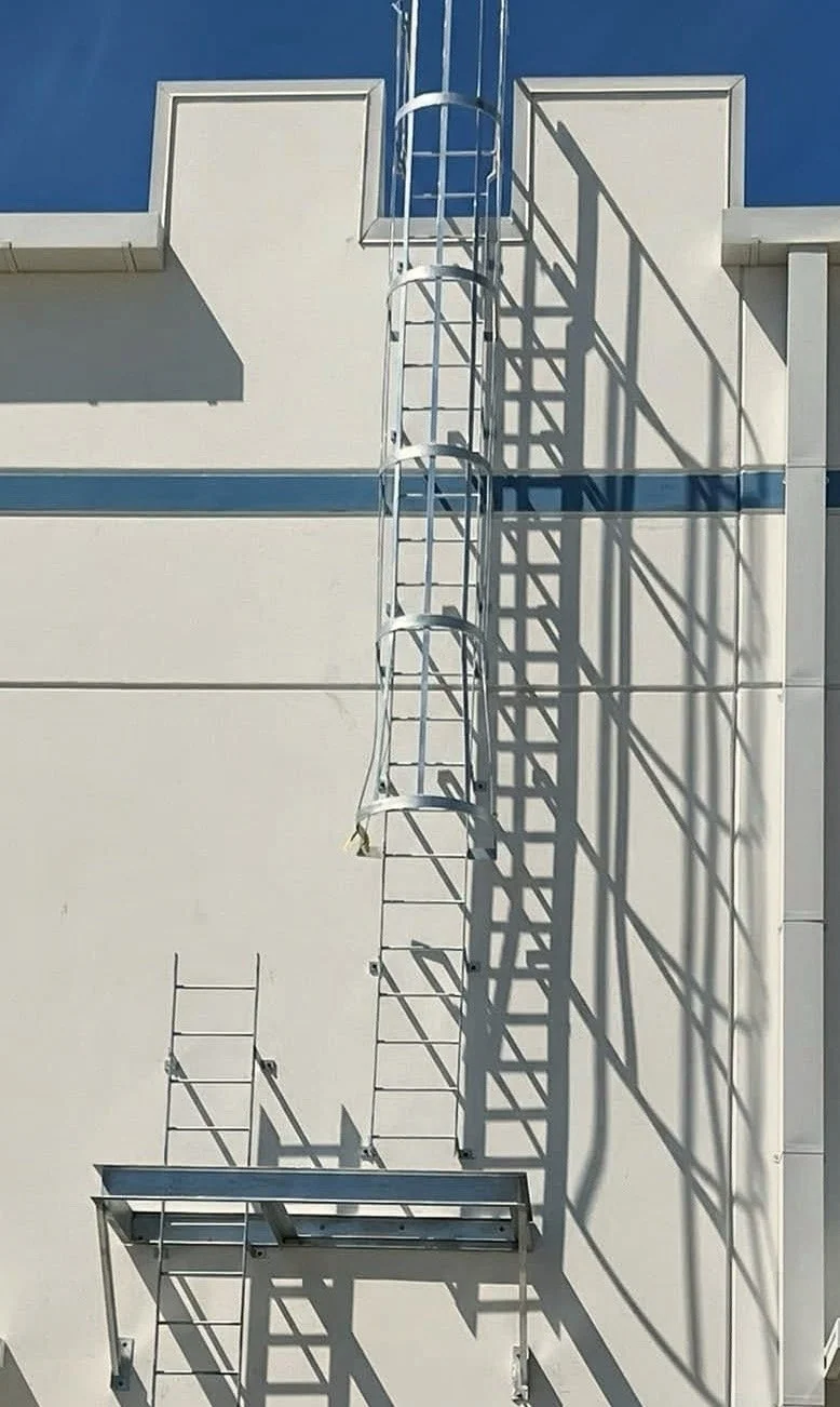 Metal fire escape ladder and platform attached to a white building, casting shadows on the wall amidst clear blue sky.