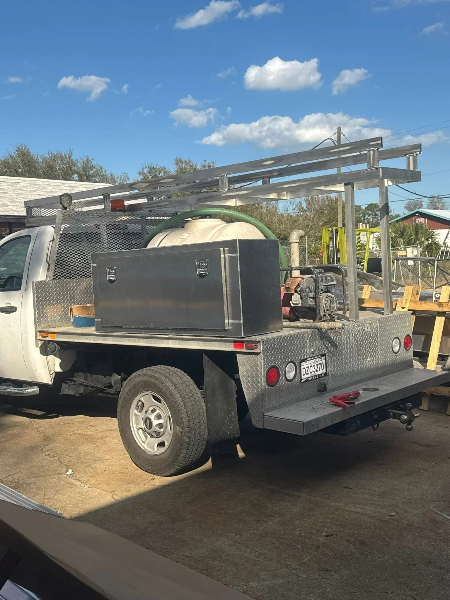 A white work truck with a metal utility bed carrying tools, equipment, and a large hose, parked on a dirt surface under a blue sky with clouds.