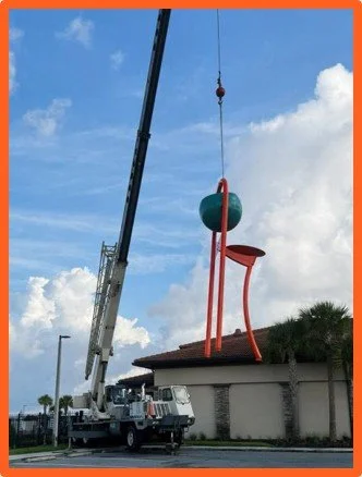 Crane lifting a large sculpture of a red chair with a curved seat and a spherical top, in front of a building with palm trees.