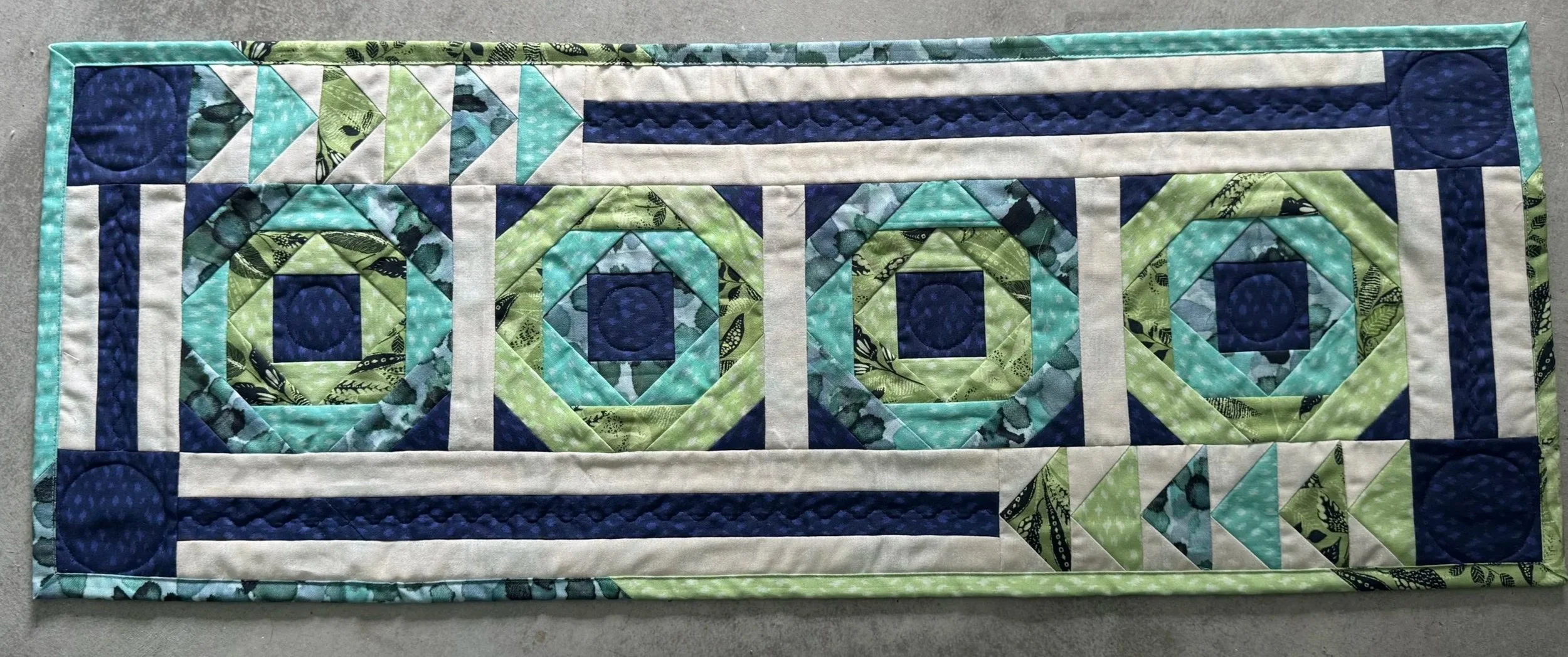 A rectangular quilted table runner with a pattern of four large, circular, and square geometric designs in shades of green, blue, and cream, bordered with a green and blue fabric.