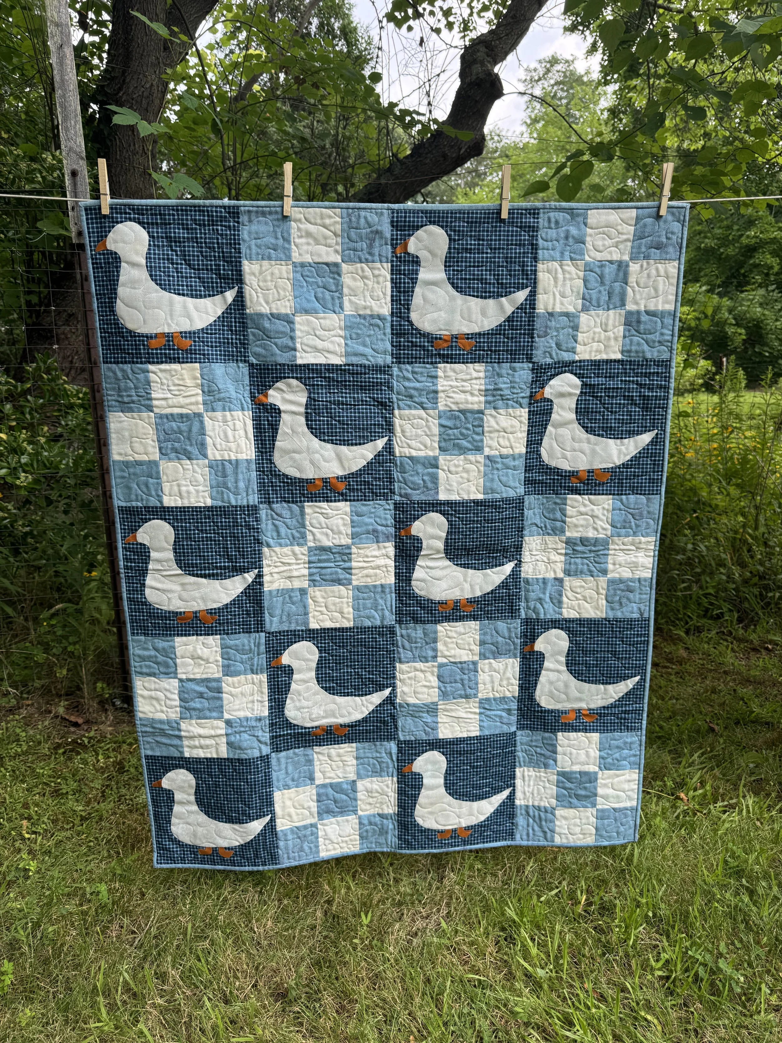 A quilt hanging on a clothesline outdoors. The quilt features a pattern of white duck appliqués with orange beaks and feet, against a background of blue checkered and blue patterned fabric. The setting includes greenery and trees.