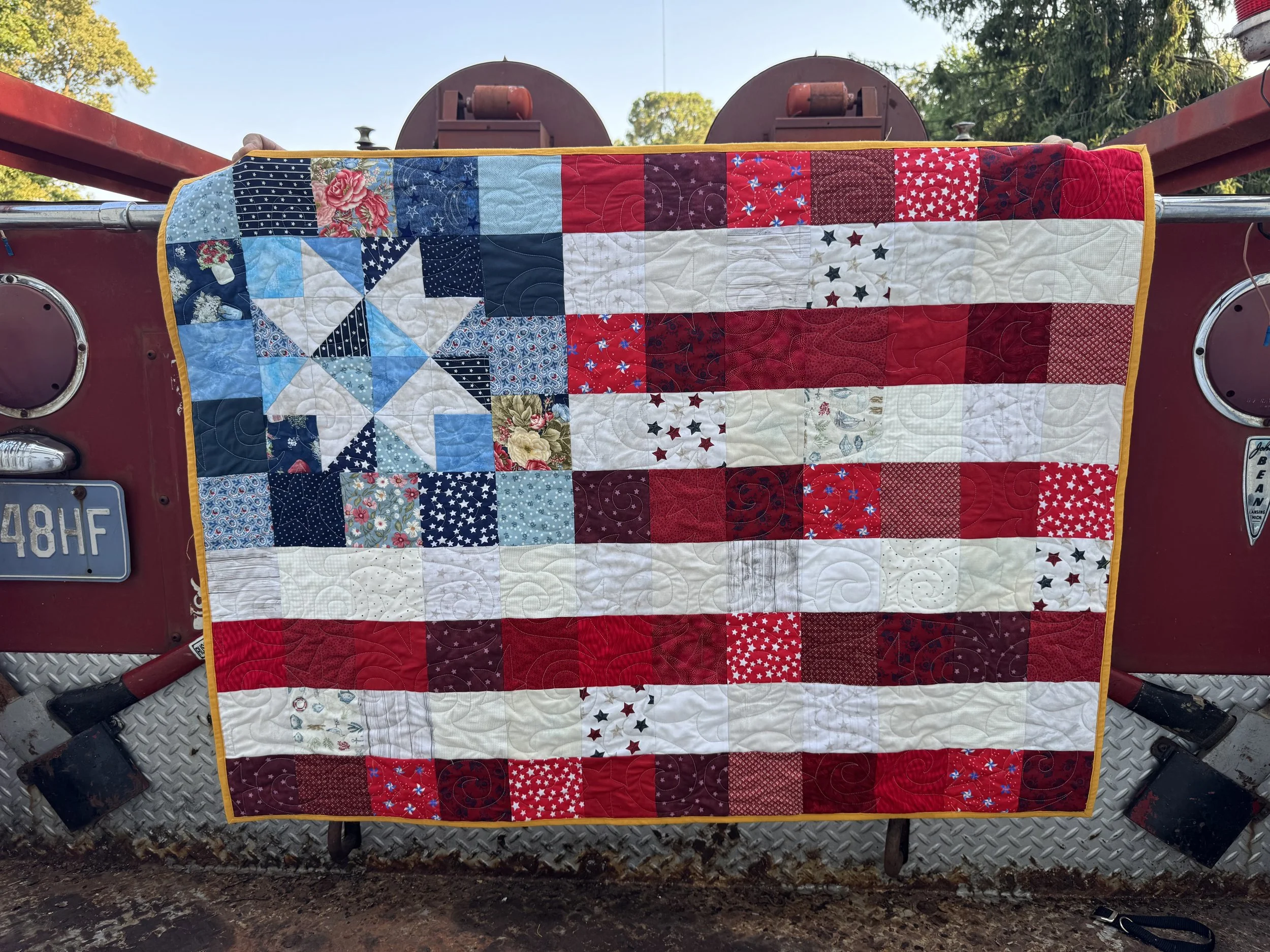 Red vintage truck with a quilt draped over the tailgate, showing a patriotic pattern of stars, stripes, and floral designs.