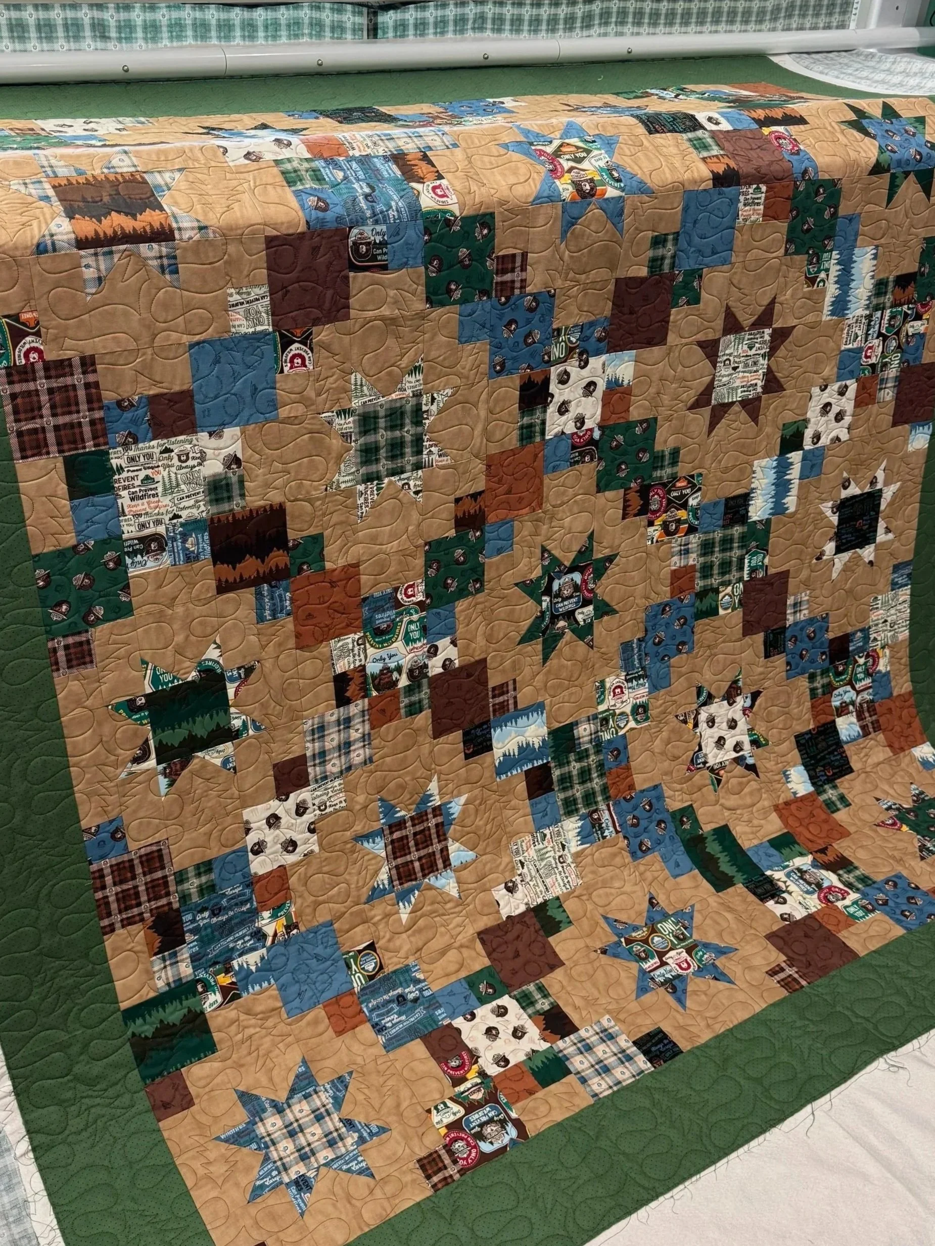 A colorful quilt with a nature and outdoor theme, featuring images of mountains, trees, and campfire scenes, arranged in star-shaped patterns on a beige background with a green border.