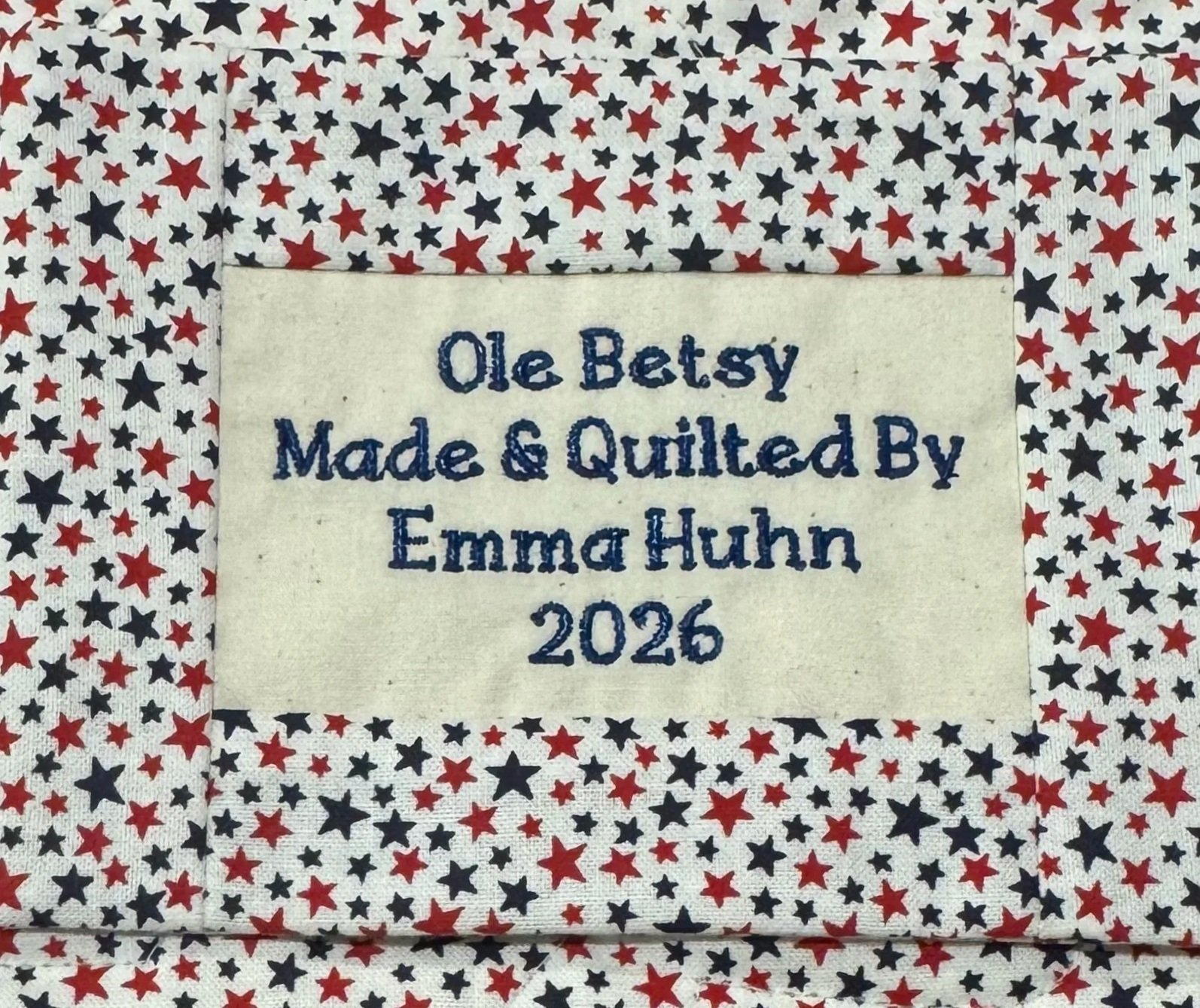 A quilt with a red, white, and blue star pattern and a label that reads, "Ole Betsy Made & Quilted By Emma Huhn 2026."