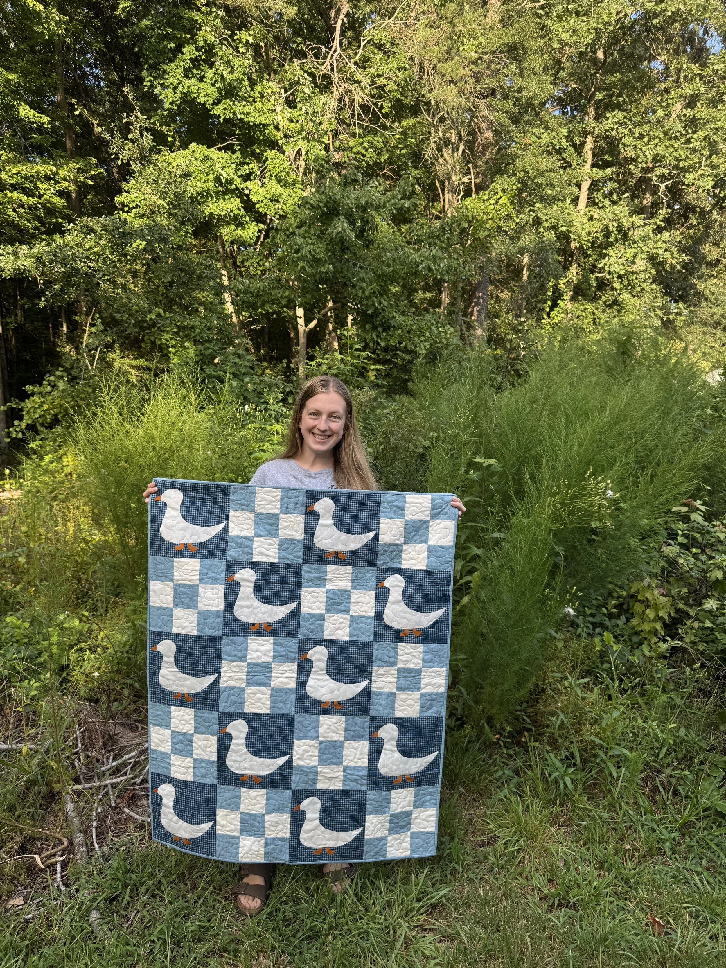 A young woman smiling and standing outdoors in a green, wooded area, holding a quilt with a duck pattern.