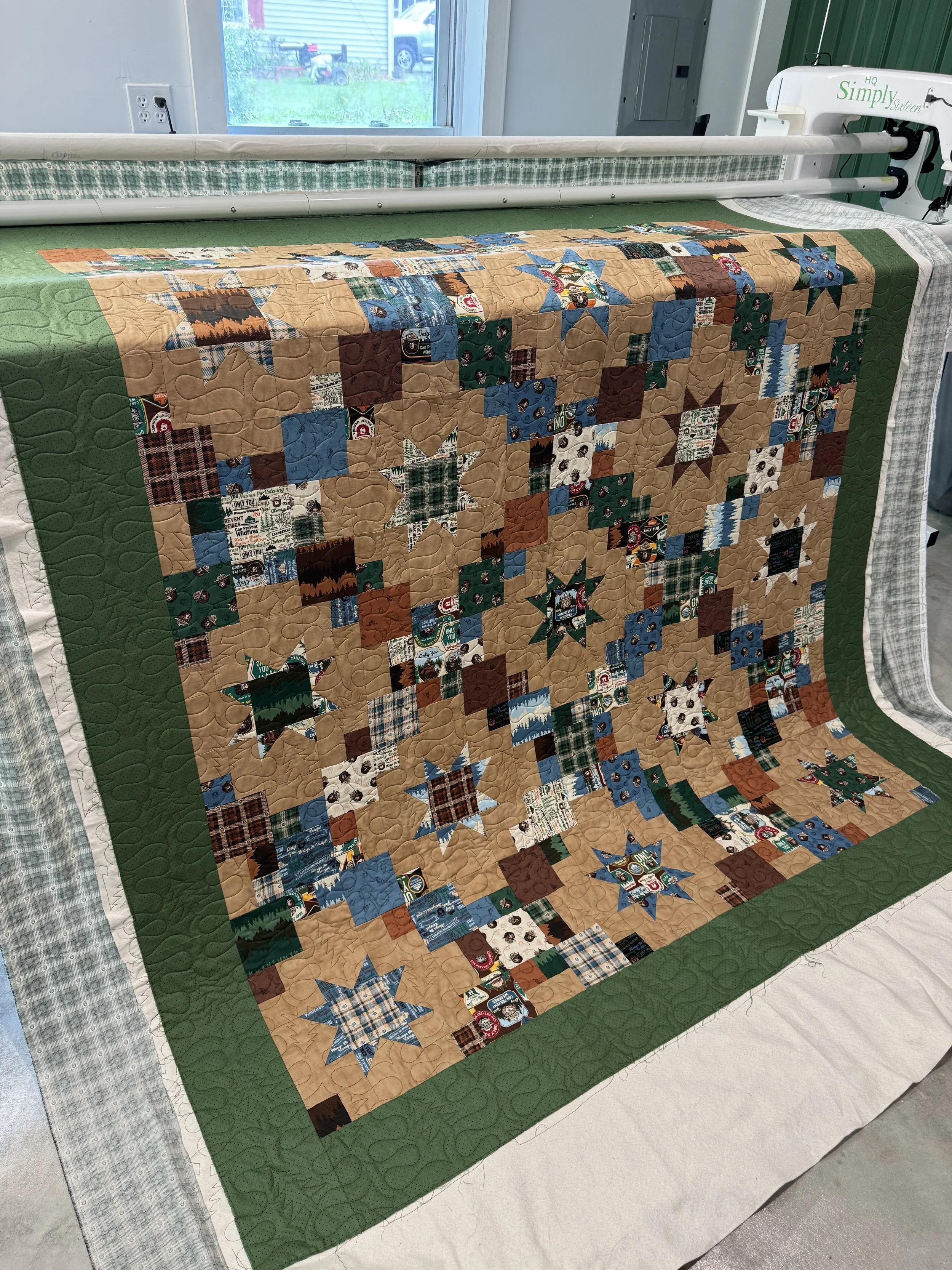 A quilt with a patchwork design featuring stars and squares, predominantly in shades of blue, brown, and plaid patterns, displayed on a bed or table, with a green border and quilted stitching visible.