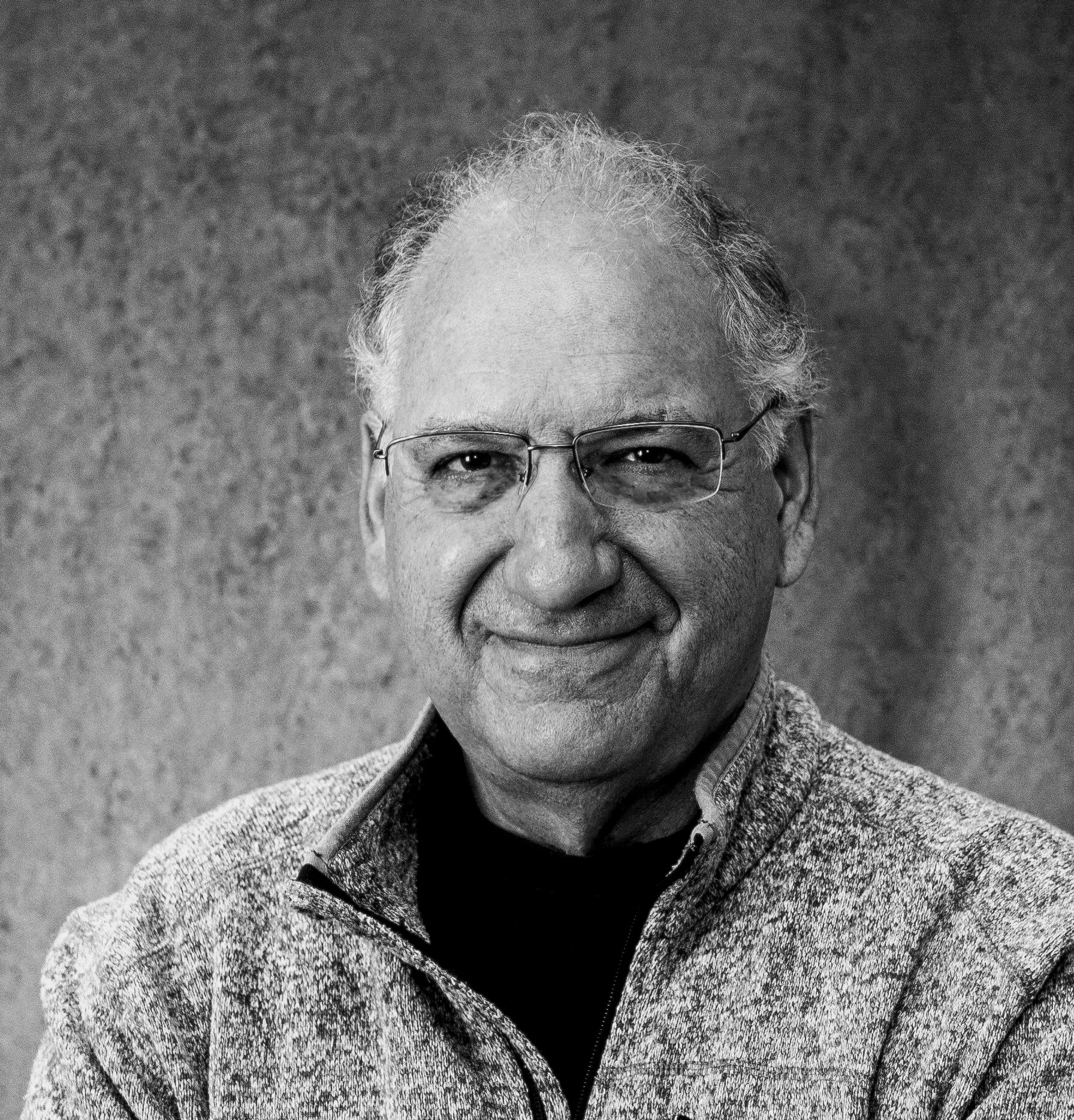 Black and white portrait of an elderly man with glasses, gray hair, and a patterned jacket, smiling gently against a plain background.