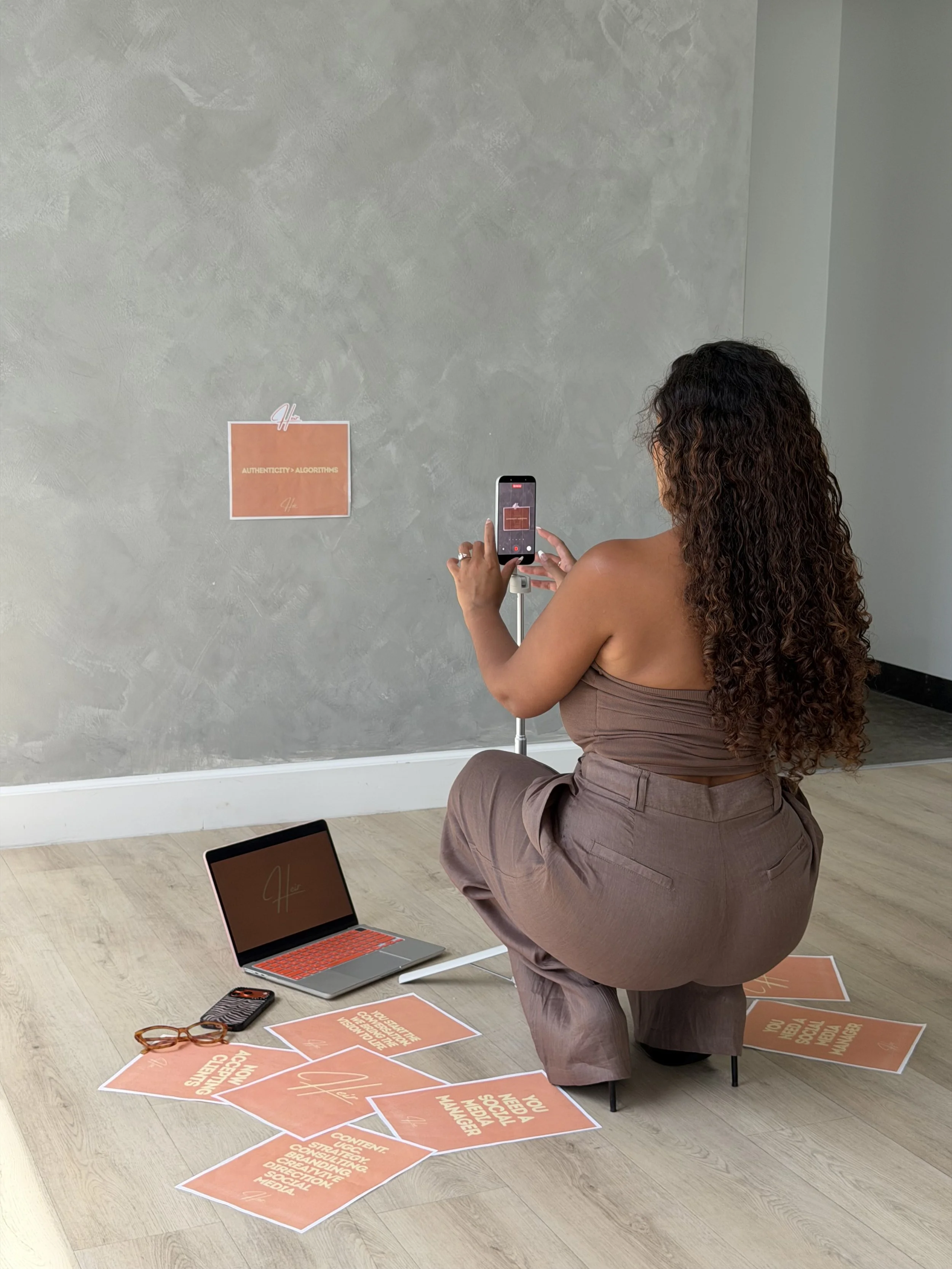 A woman with curly hair kneeling on one knee on a wooden floor, taking a photo with her smartphone of a sign on the wall that reads 'Authenticity > Algorithms'. Several orange signs with white text are scattered on the floor, with some on a table. The signs include phrases like 'Content strategy, branding, digital marketing, social media', and other related phrases.
