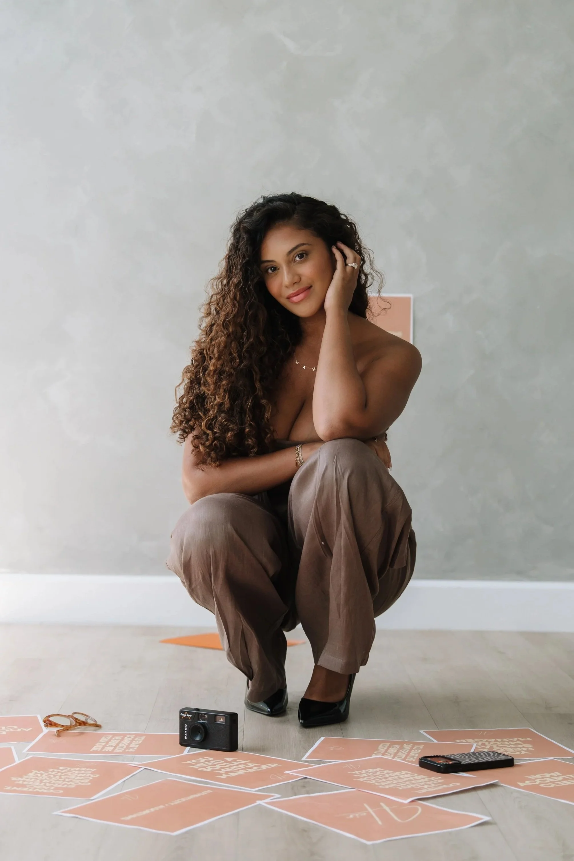 Woman with long curly hair, kneeling with one knee up, in front of a plain wall. Papers with writing scattered on the floor, a camera, smartphone, and pair of glasses around her. She is touching her face, smiling softly, dressed casually.