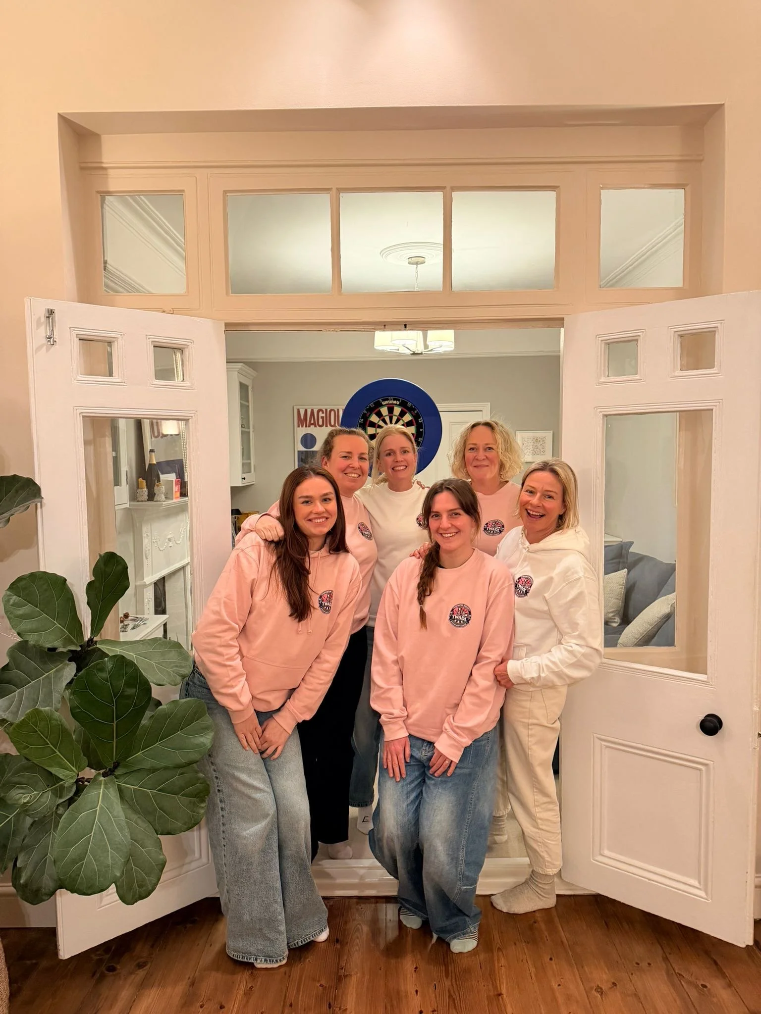 A group of seven women standing together inside a house, smiling for a photo. They are standing in a doorway with open doors and a dartboard is visible in the background. The women are wearing casual clothing, some with matching pink hoodies, and the room has wooden floors and a plant in the foreground.