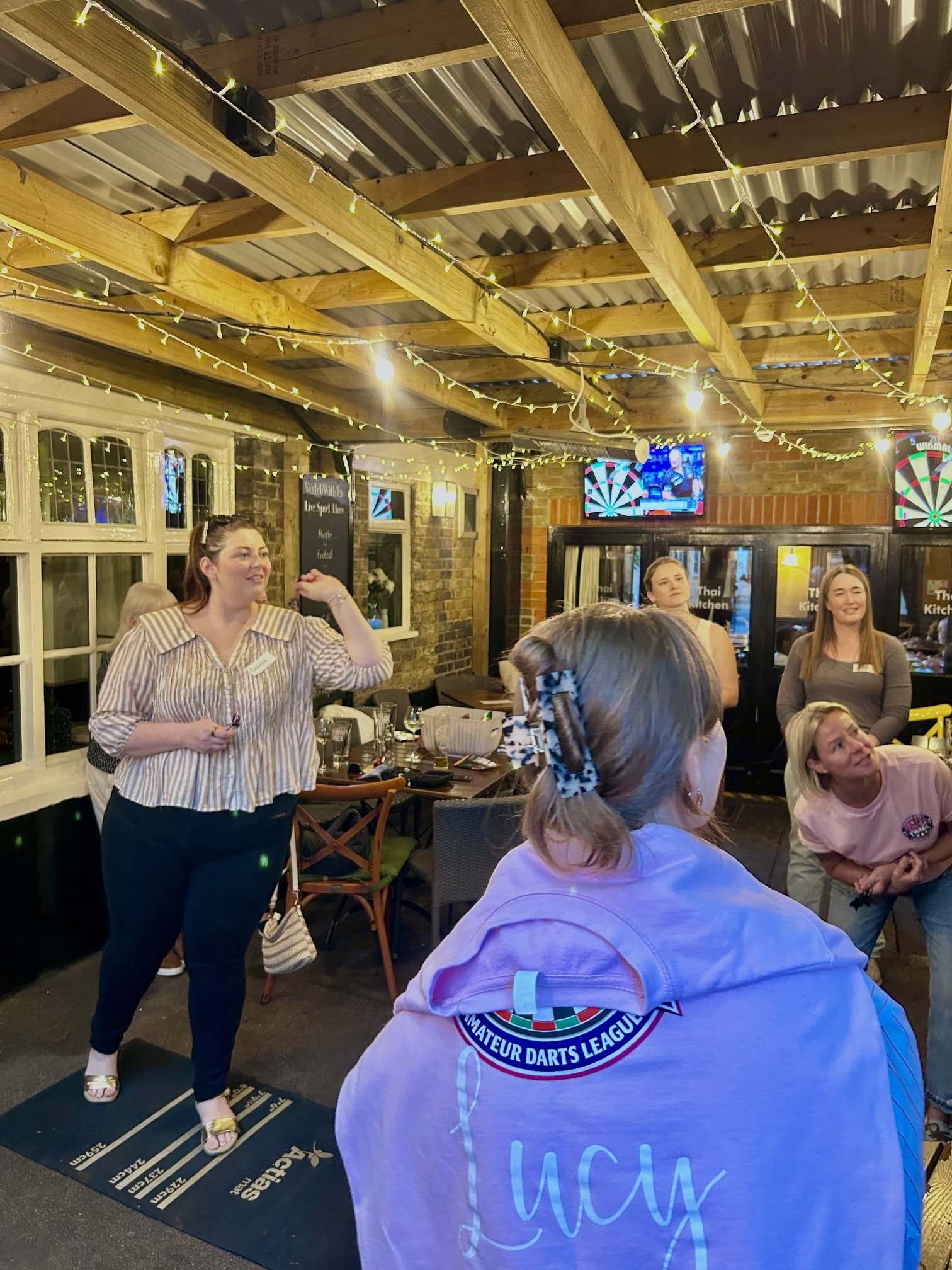 Group of women gathered indoors at a social event, some smiling and talking, with a woman in a striped shirt standing near a dartboard, and a dartboard game on the wall. The ceiling is decorated with string lights and wooden beams, and there is a tel
