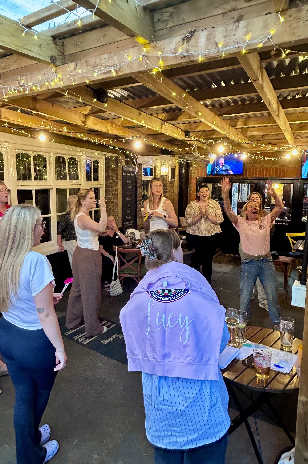 Women and a girl gathered in a cozy pub or restaurant, some cheering and watching a TV, with warm wooden ceiling, string lights, and brick walls.