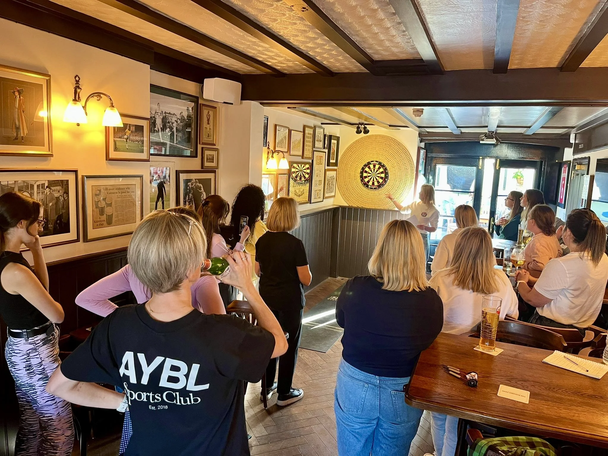 A diverse group of people gathered in a pub or bar, watching a dart game on the wall. A woman is standing and pointing at the dartboard, possibly explaining the game. The room has framed pictures and sports memorabilia on the walls, with a warm, invi