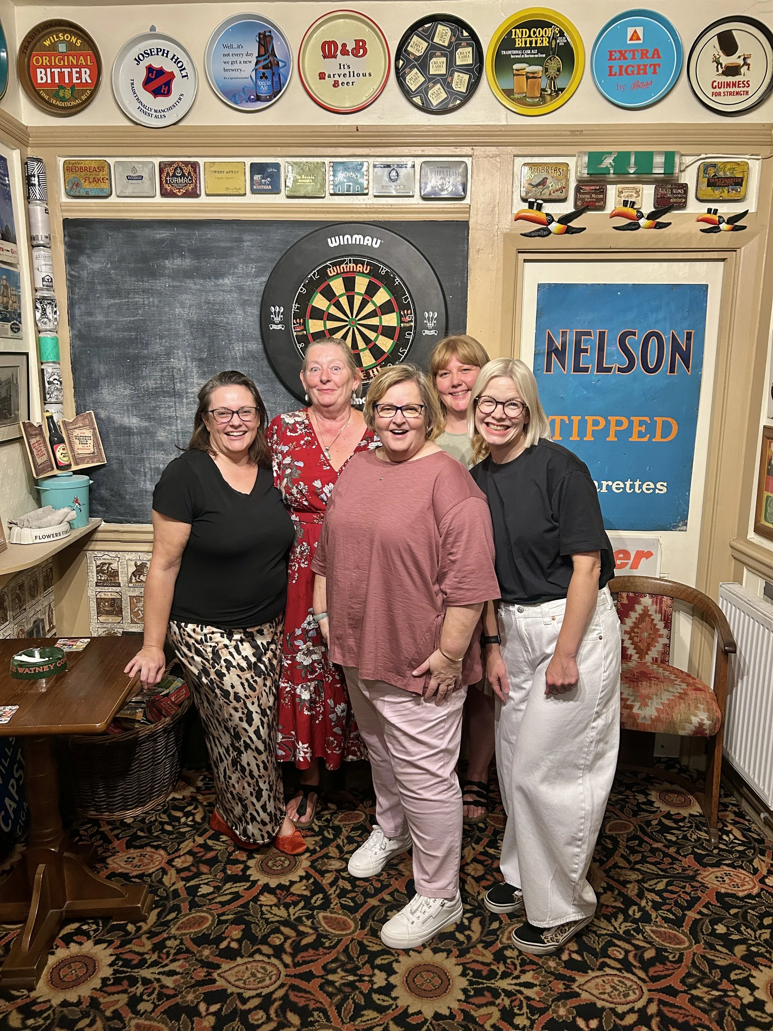 Five women smiling and posing together in a pub or bar. The background features a dartboard on the wall, vintage beer signs, and a patterned carpet.
