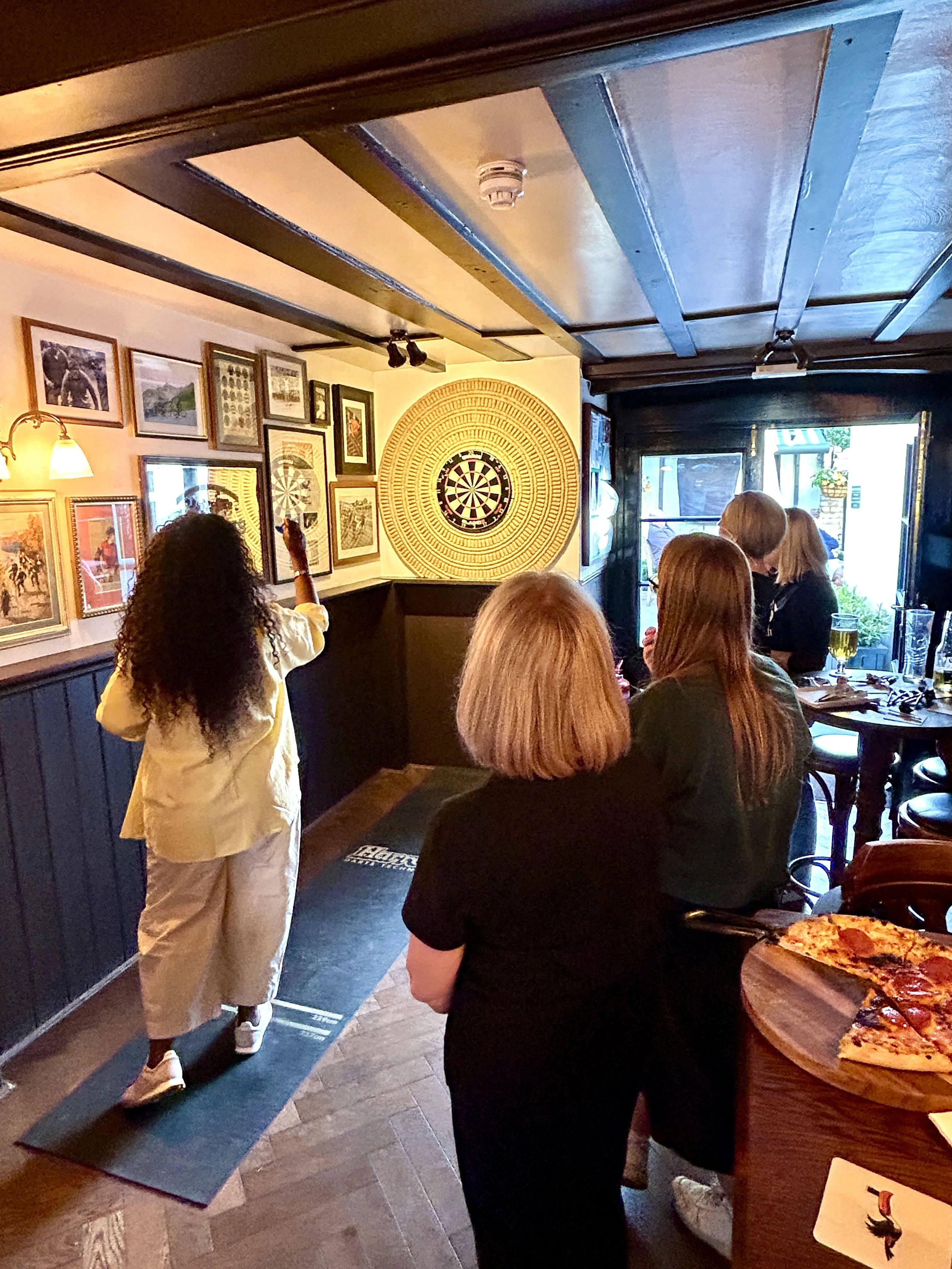 A woman with curly hair and a yellow shirt is playing darts in a cozy, decorated room filled with framed pictures and a large dartboard on the wall. Several people are gathered, some sitting and some standing, around a table with pizza and drinks.