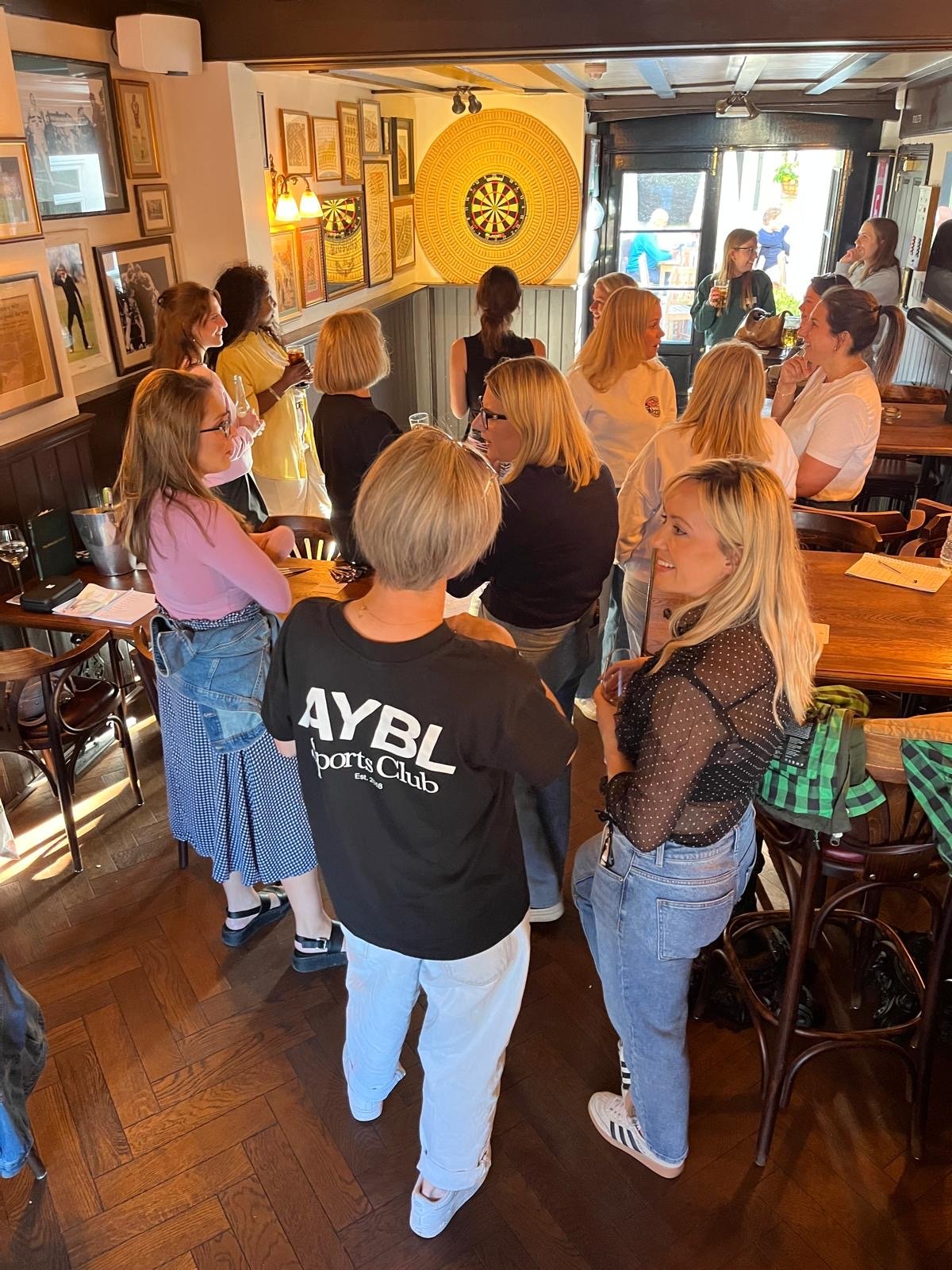 A group of women and young women gathered in a pub, talking and socializing near a dartboard on the wall, with some holding drinks.